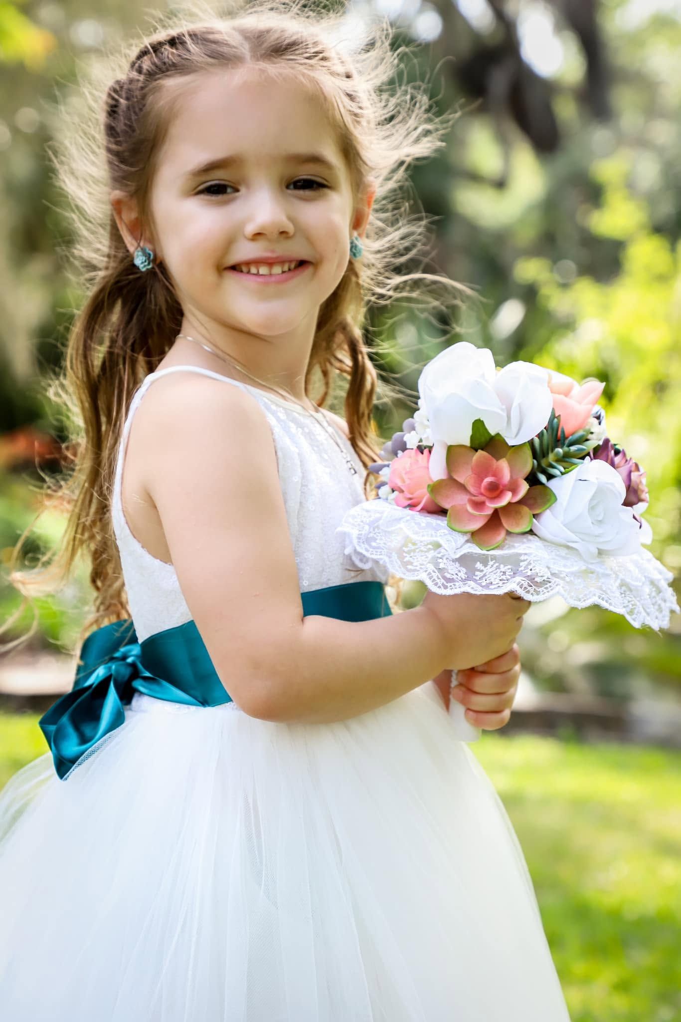 A smiling flower girl in a white dress with teal bow holds a floral bouquet outdoors in Gainesville Florida.