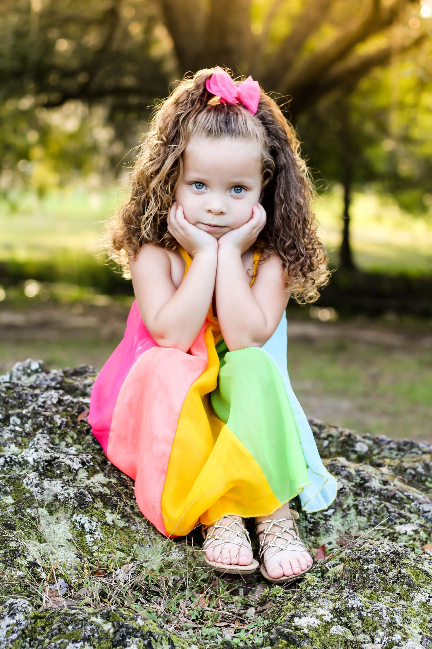 Portrait of Curly-haired little girl in rainbow dress sitting on rock with hands on cheeks in sunny park.