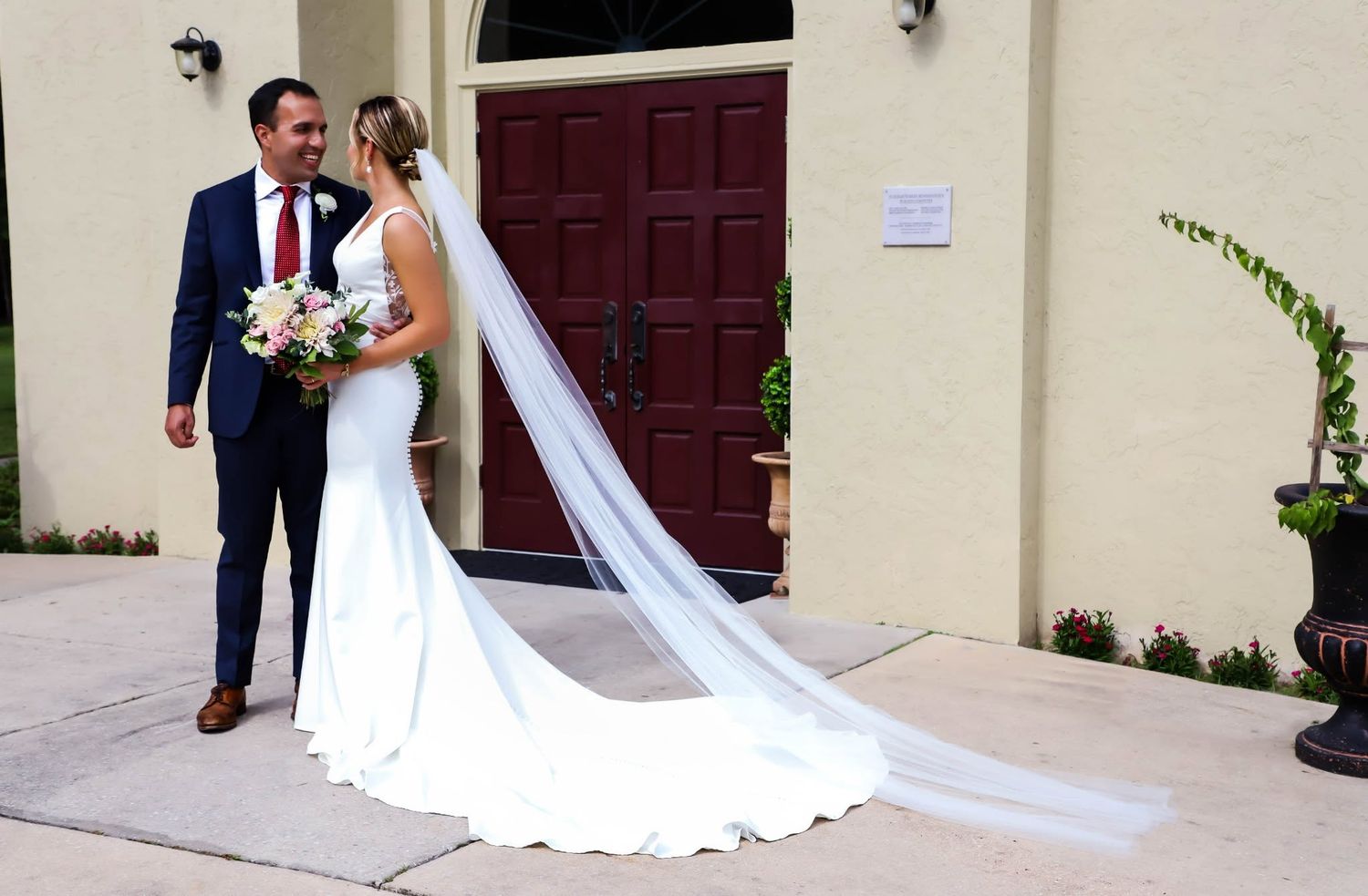 Bride and groom standing together outside elegant building with long cathedral veil trailing behind.