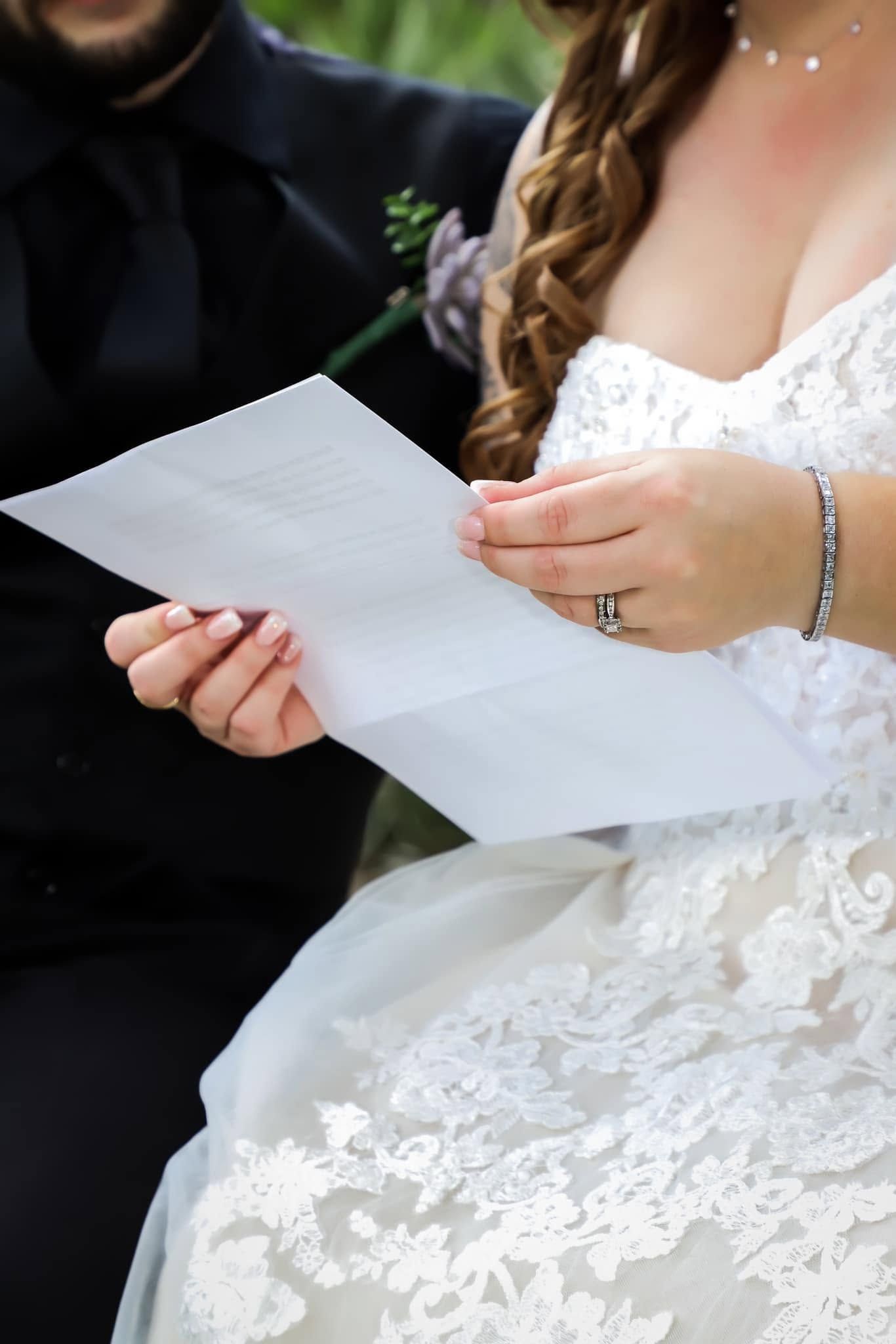 Bride in lace wedding gown reads vows from paper while groom in black suit sits beside her during ceremony.