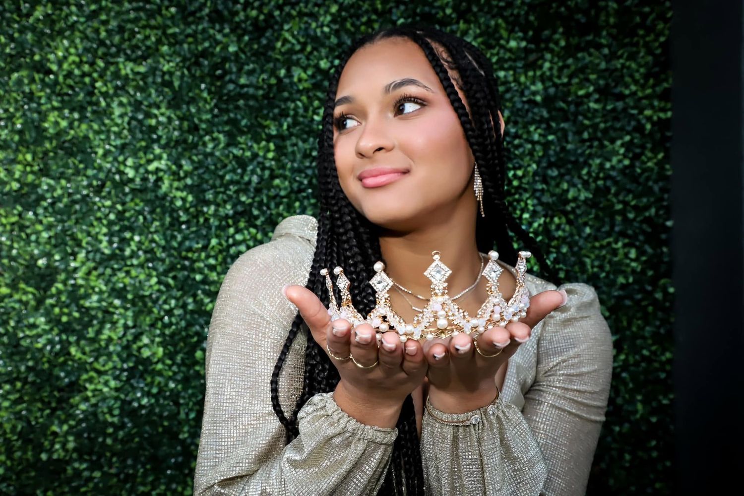 Young woman with long braids holding small decorative crown in outstretched hands against green wall.