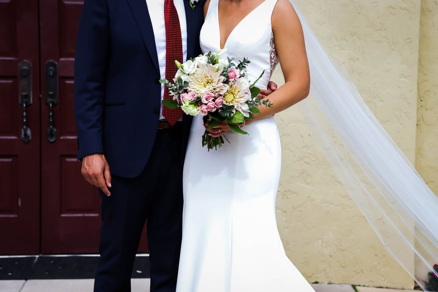 Bride holding a colorful bouquet poses with groom in navy suit outside a brick building.
