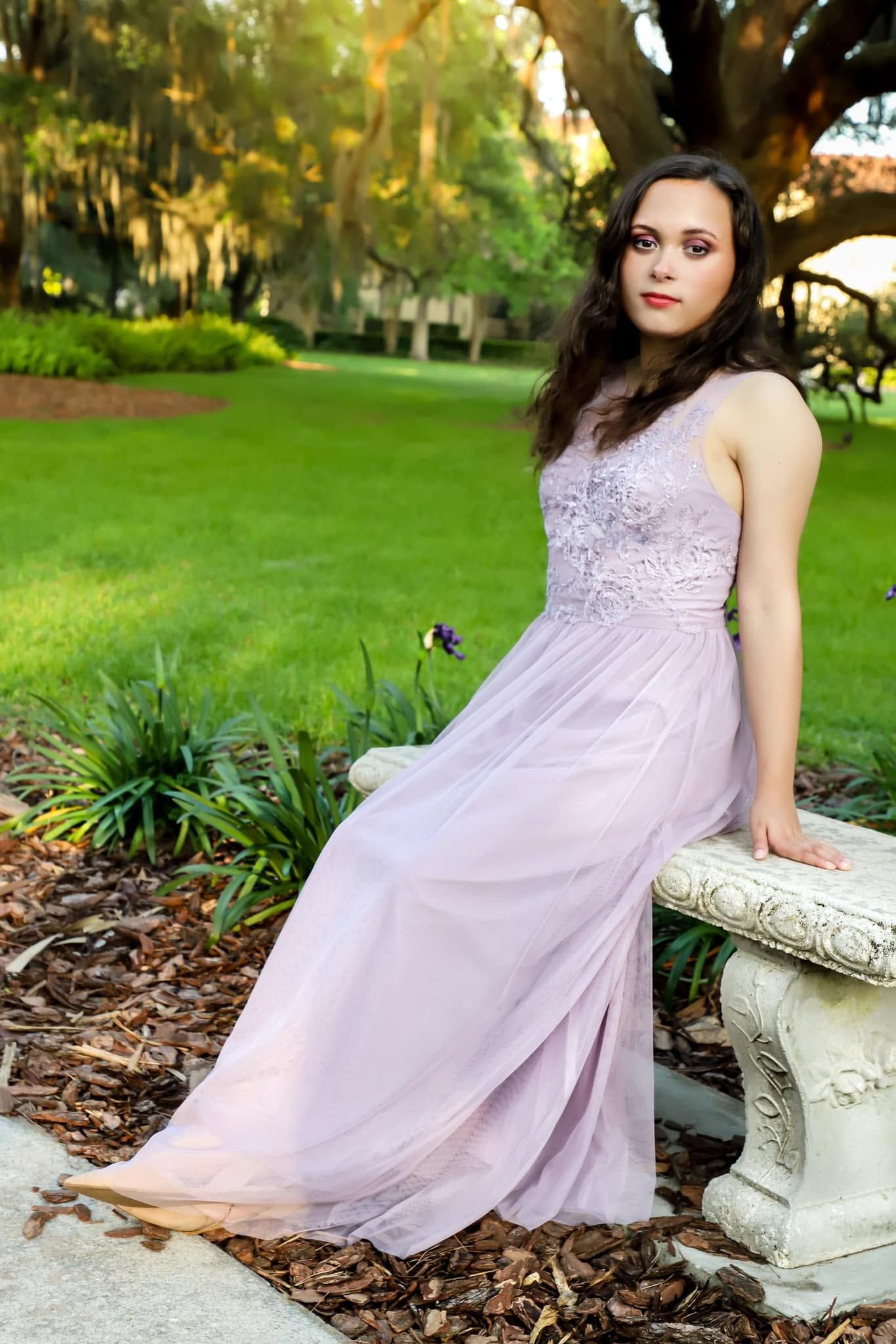 Young woman in flowing lavender gown poses gracefully beside a stone bench in a lush green garden setting.
