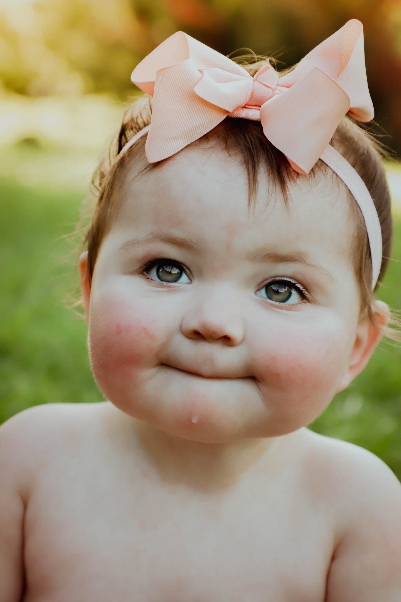 Adorable baby girl with rosy cheeks wearing a pink bow headband smiling outdoors.