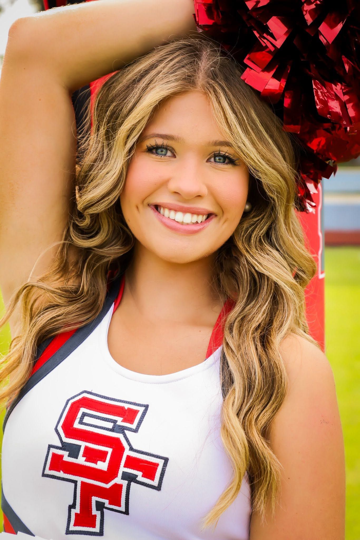 Smiling blonde cheerleader in SF uniform holds red pom pom at outdoor sports field portrait.