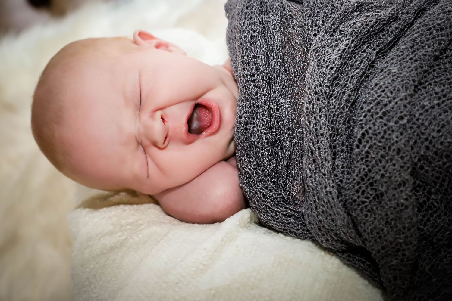 A swaddled newborn baby yawns while wrapped snugly in a soft gray knit blanket on a white surface.