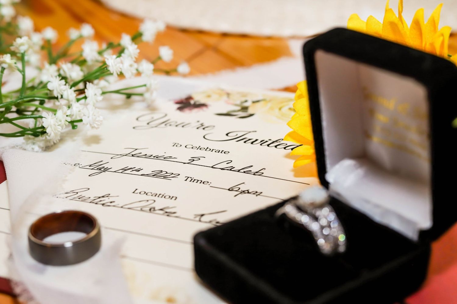 Wedding invitation with diamond engagement ring in black velvet box, wedding bands, and white flowers on table.