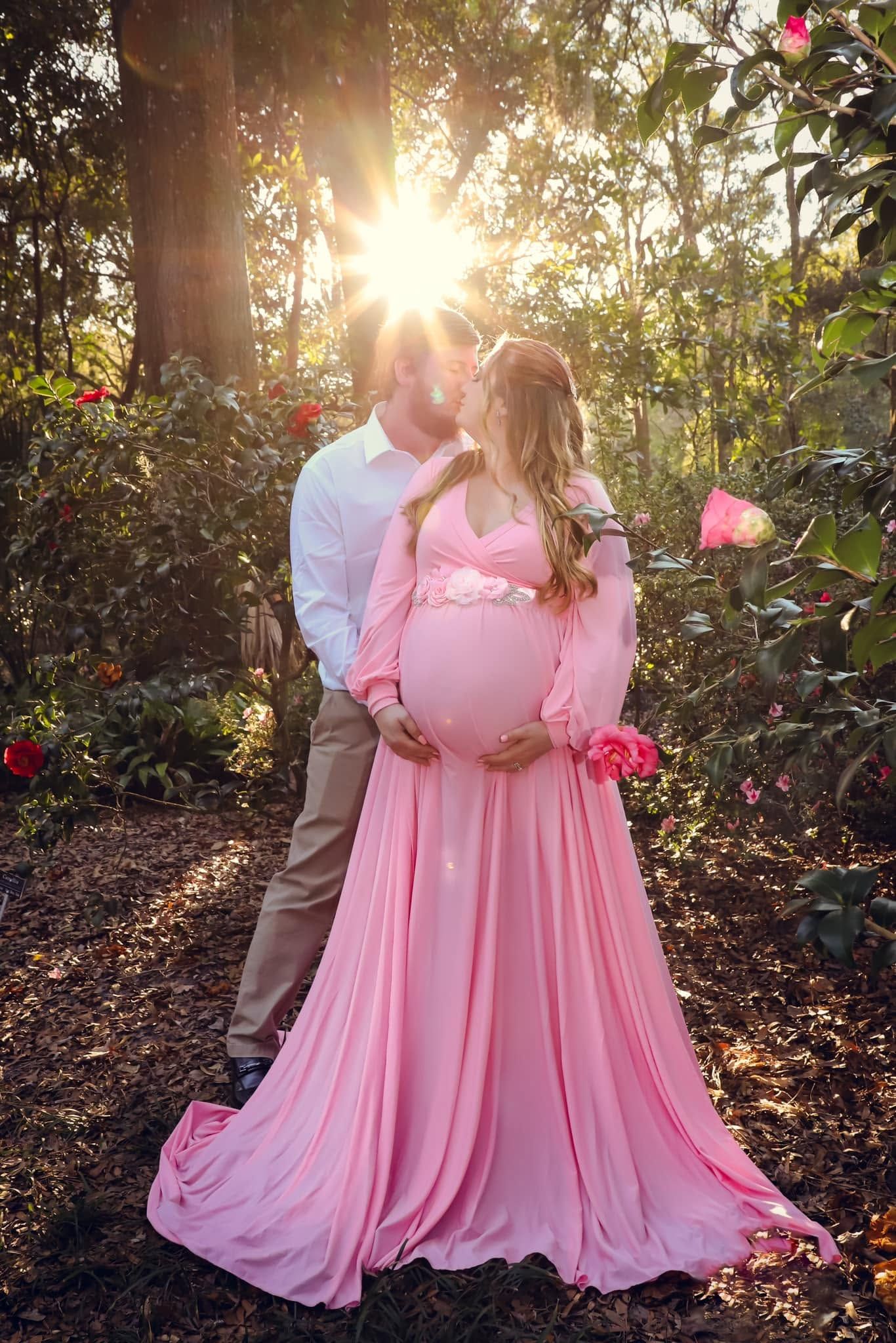 Pregnant couple kiss at sunset in a garden, with the woman in a flowing pink maternity gown in Gainesville, Florida.
