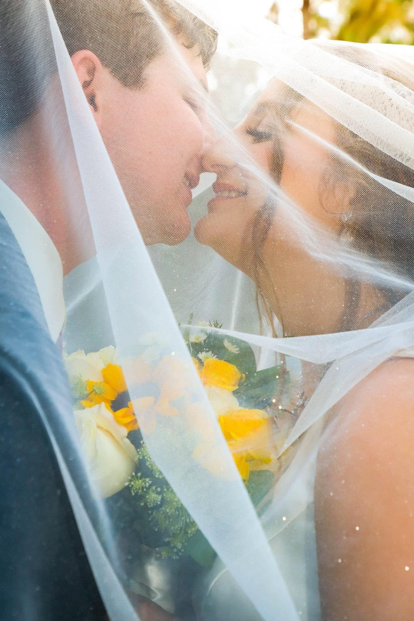 Portrait of Bride and groom sharing intimate kiss under flowing white veil, bride holding yellow rose bouquet.