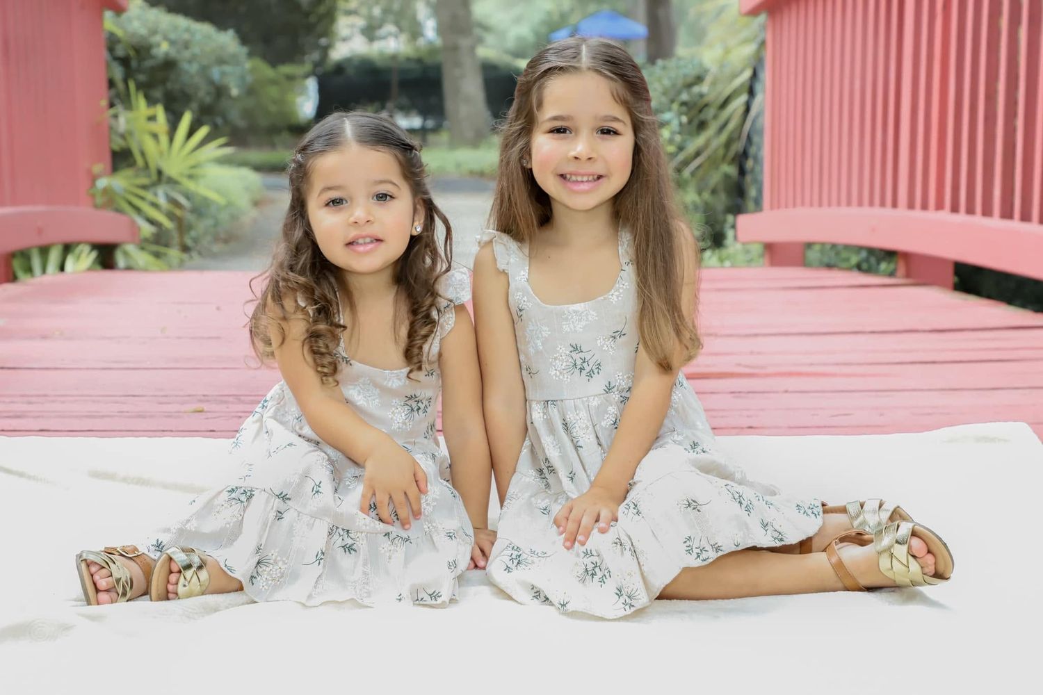 Two young girls in matching floral dresses sit together smiling on a white surface near a red bench outdoors.