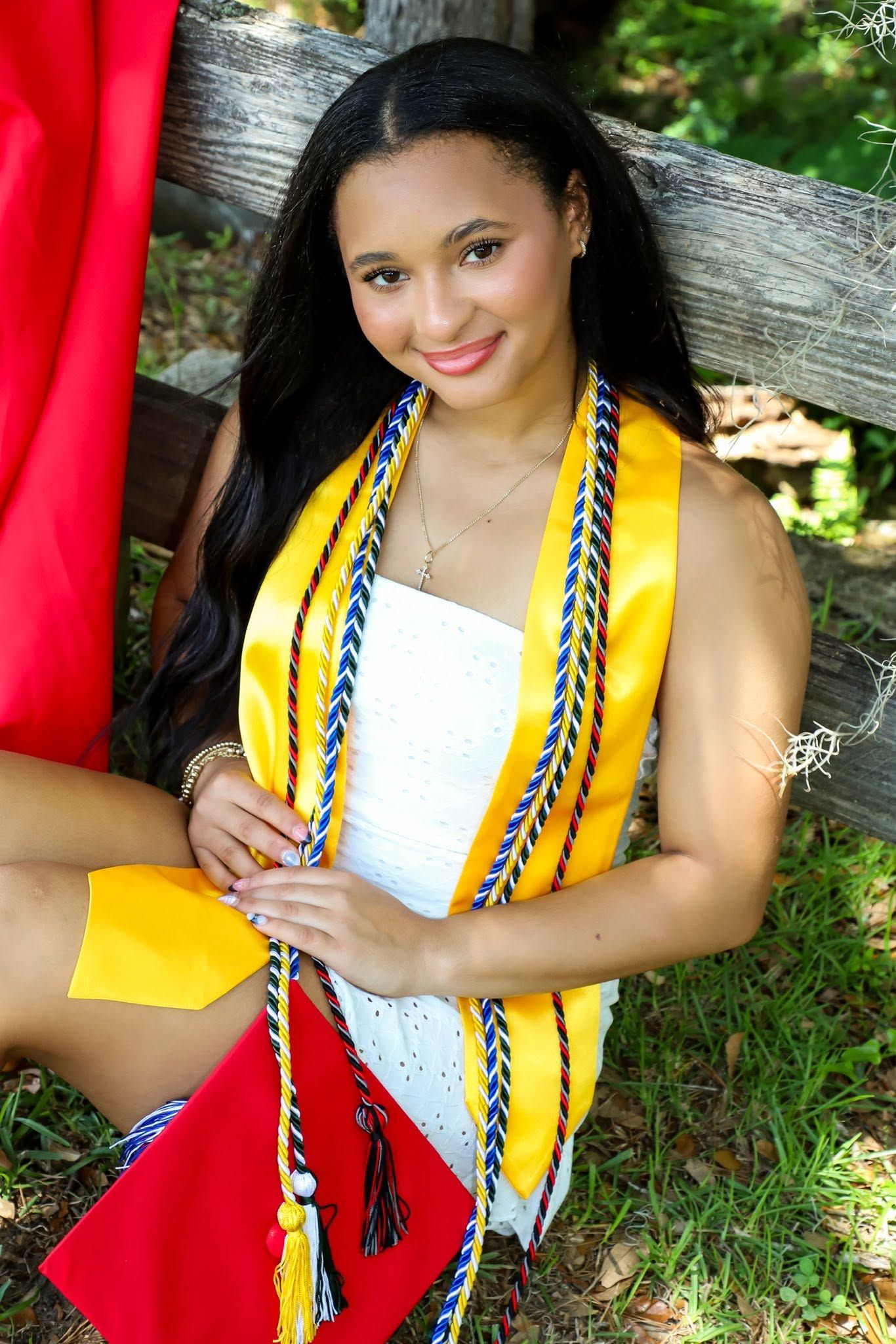 Graduation portrait of a smiling graduate in a white dress with a gold stole and colorful cords outdoors in Alachua, Florida.