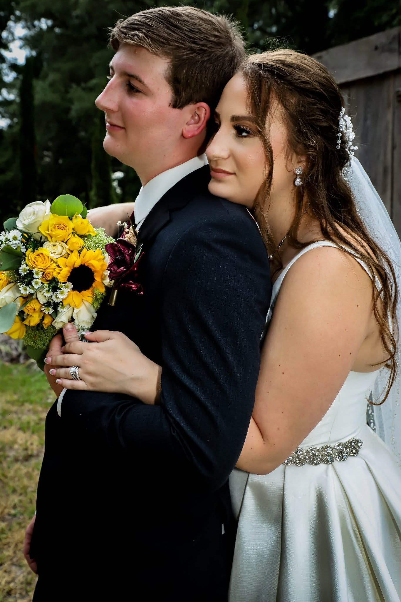 Bride and groom embracing back-to-back holding a sunflower wedding bouquet outdoors.