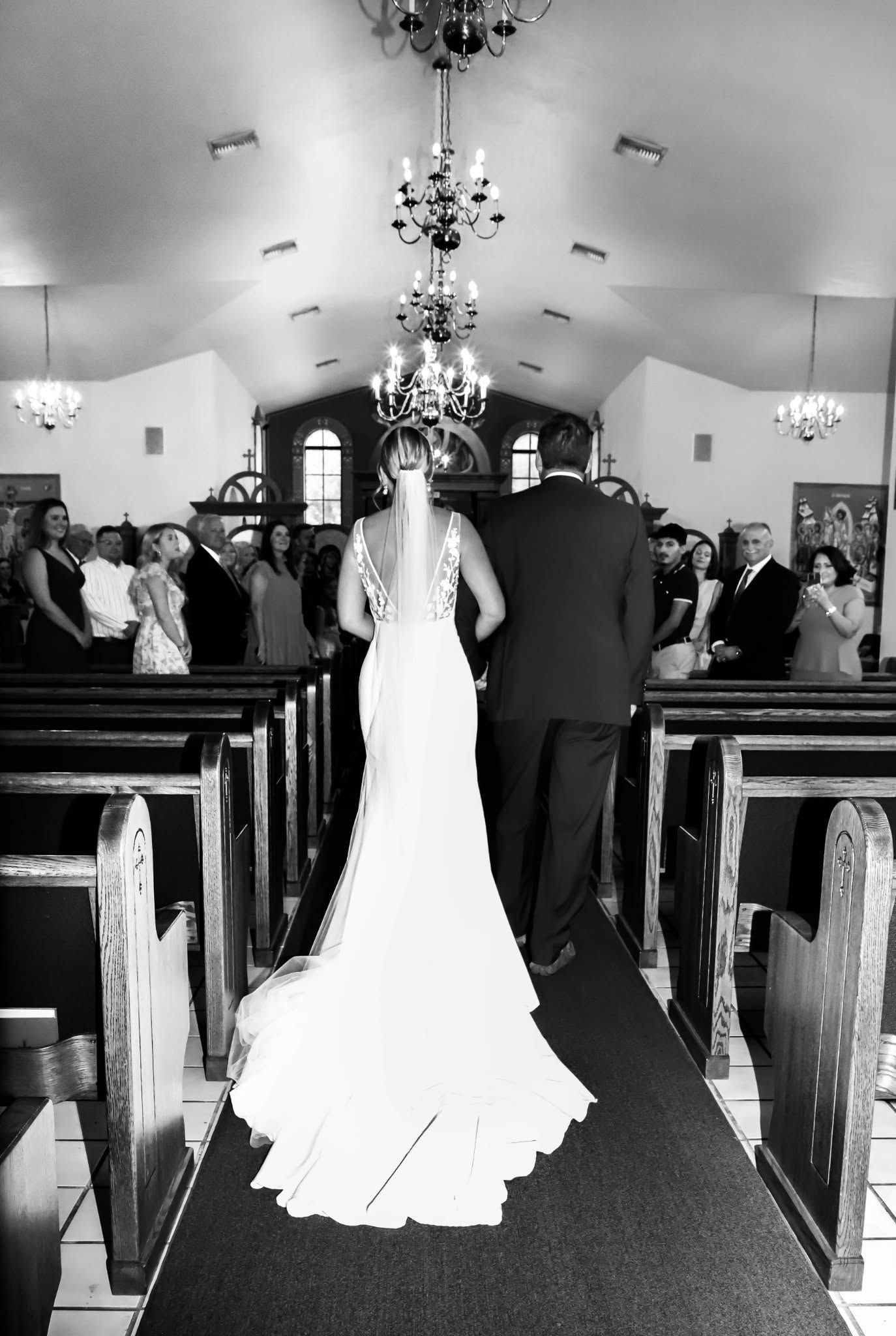 Black and white photo of bride walking down church aisle with chandelier and wedding guests.
