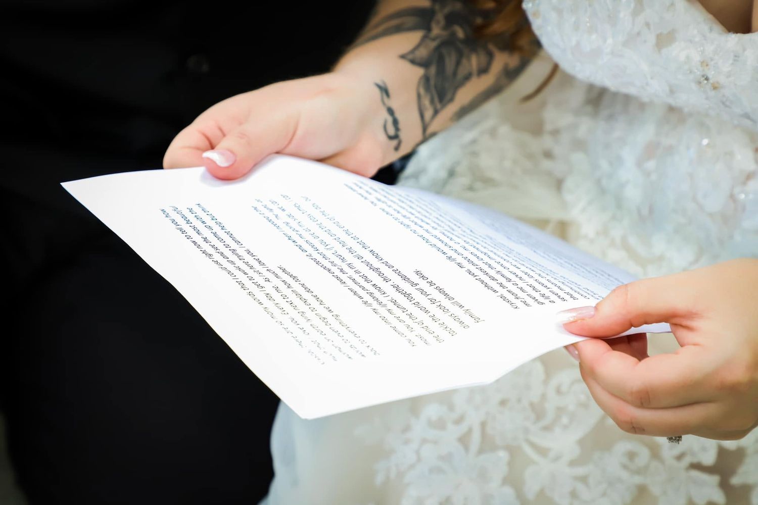 Close-up of tattooed groom and bride in lace dress holding wedding vow papers during their ceremony.