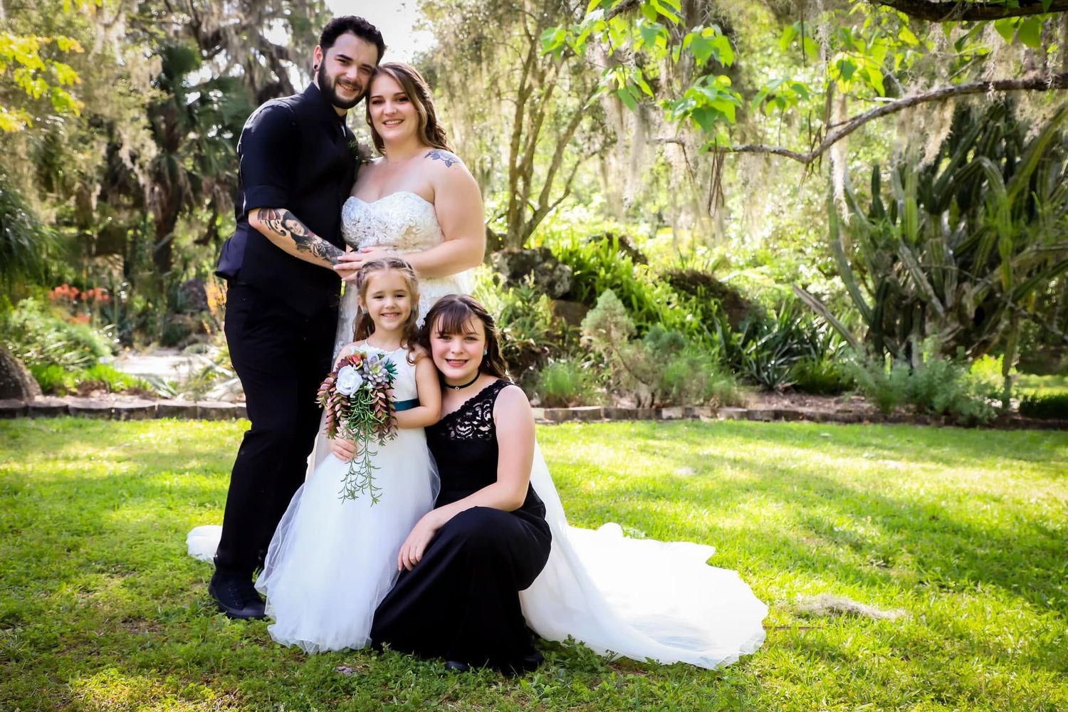Wedding family portrait with tattooed groom, bride in white gown, flower girl, and woman in black dress outdoors.
