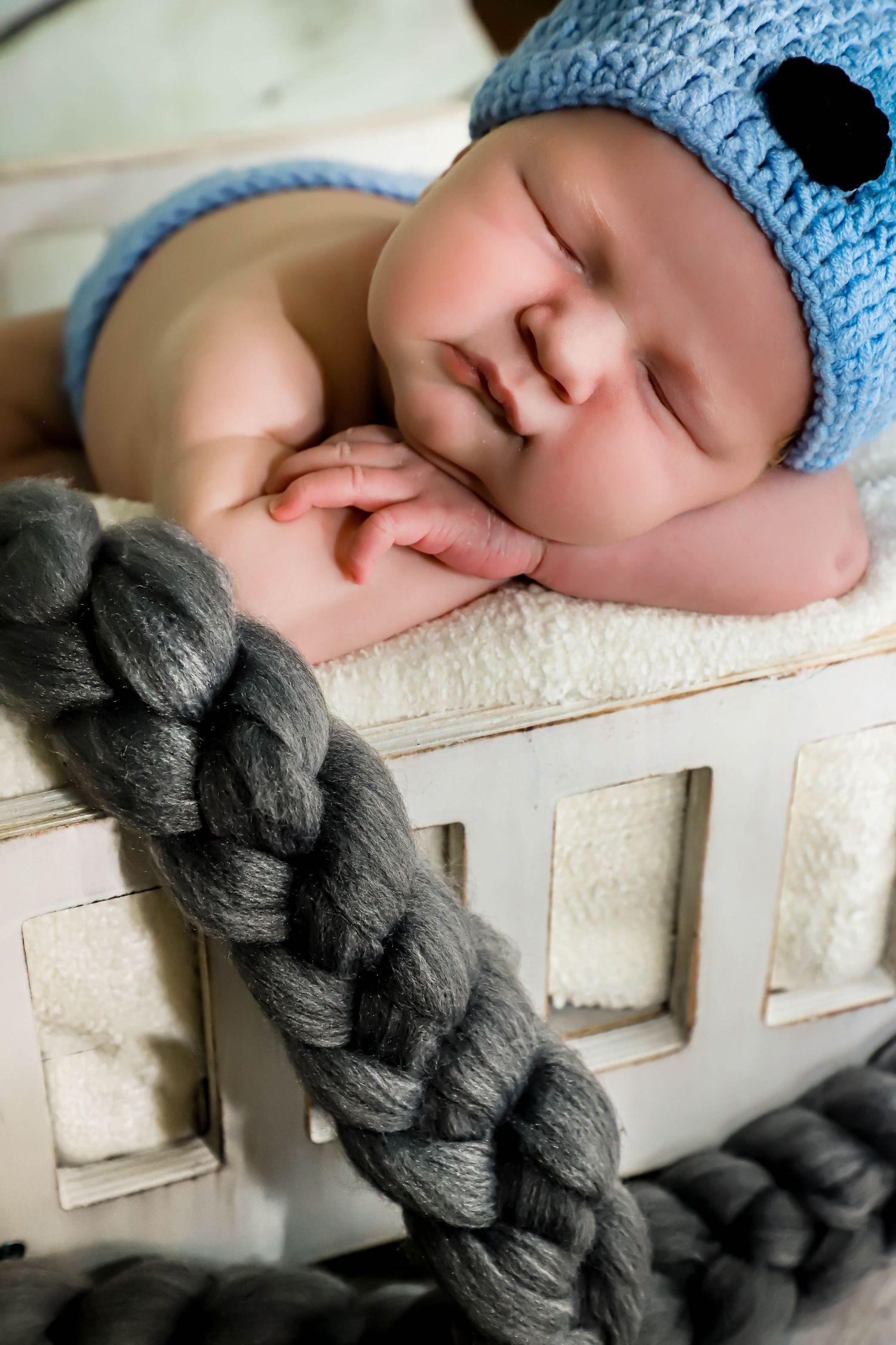 Sleeping newborn baby in blue knit hat resting in white wooden crib with gray braid.