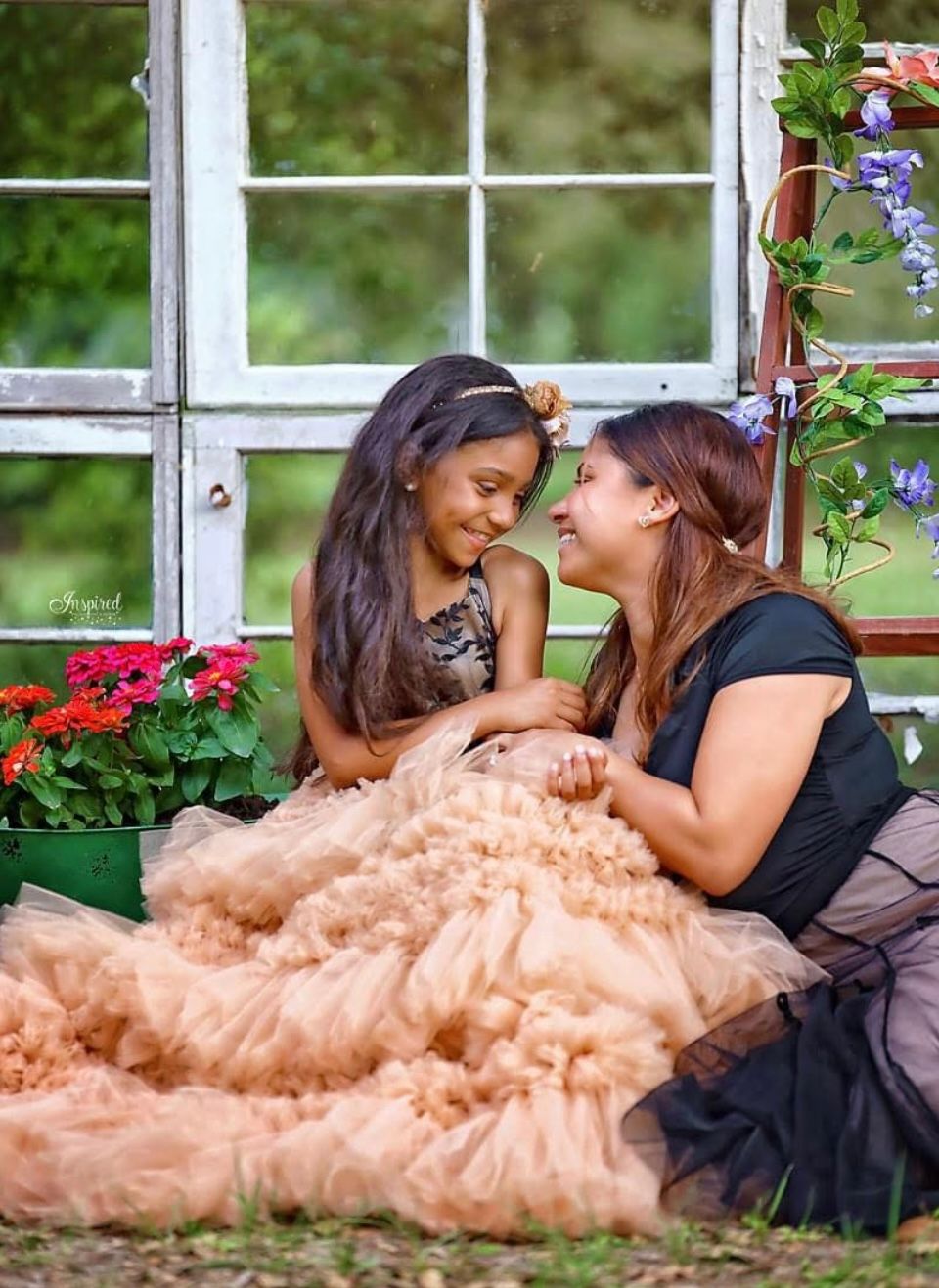 Mother and daughter sharing a joyful moment outdoors, girl wearing peach tulle gown near vintage window.