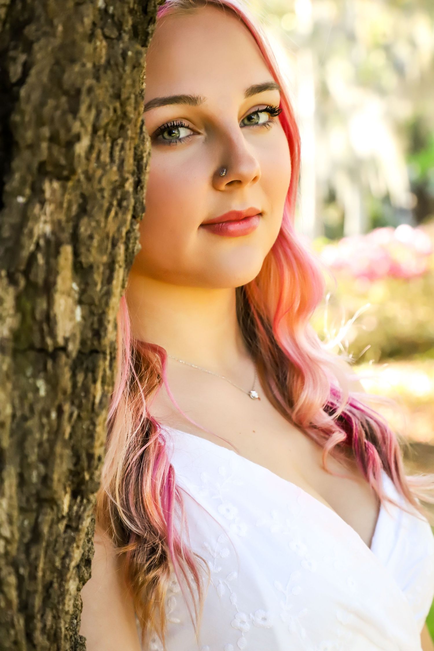 Young woman with pink highlighted hair leaning against a tree wearing a white dress outdoors.