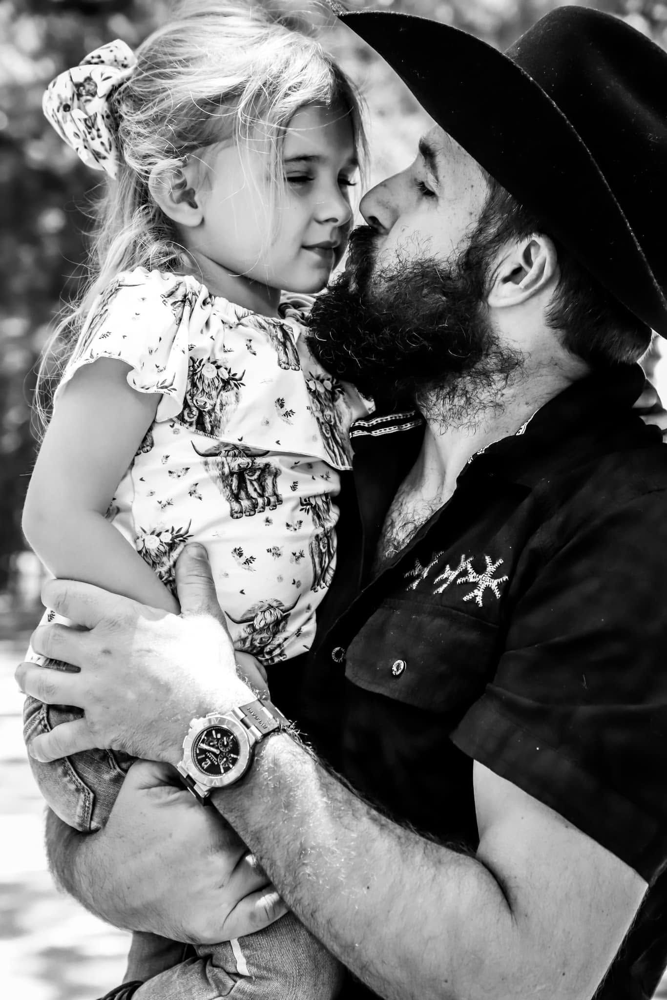 Black and white photo of bearded man in cowboy hat tenderly holding young girl.