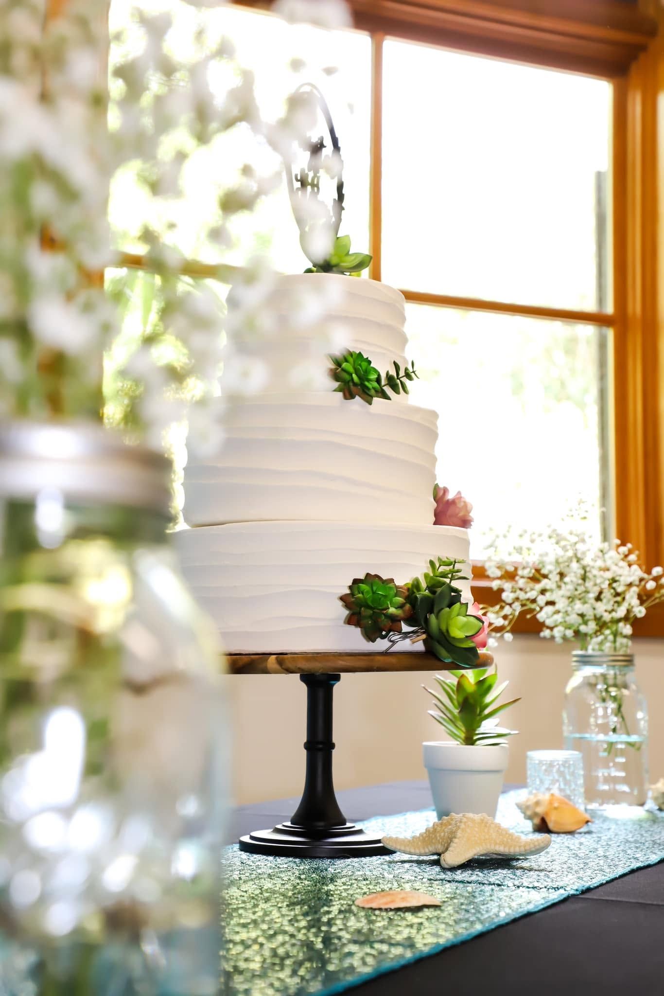 Elegant three-tier white wedding cake decorated with succulents on a dark pedestal stand near a sunny window.