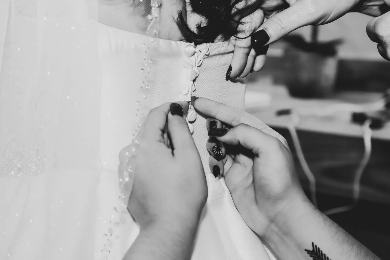 Black and white close-up of bridesmaids buttoning the back of a lace wedding dress before the ceremony.