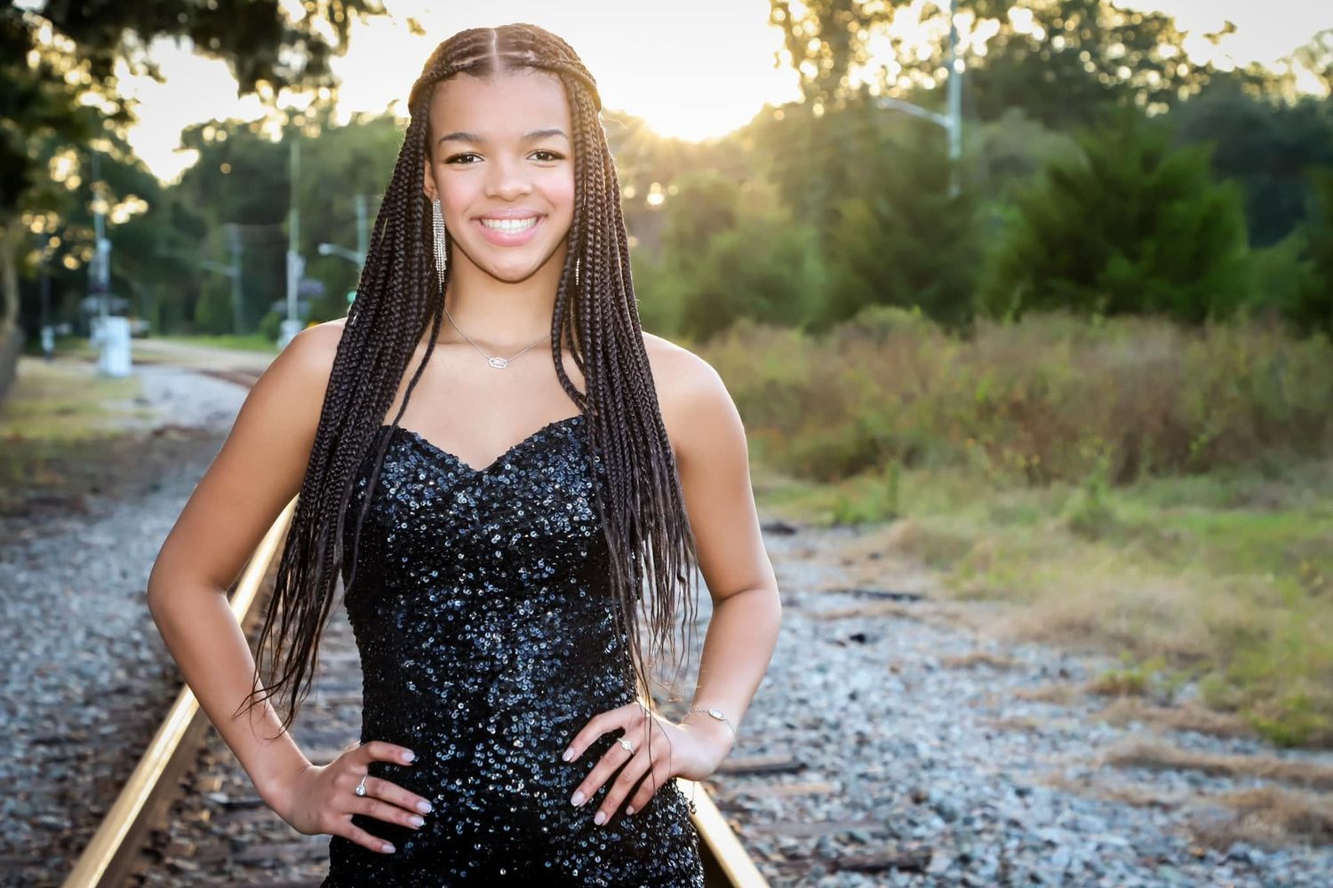 Teen girl with braids in a black sequin dress posing on railroad tracks at sunset.