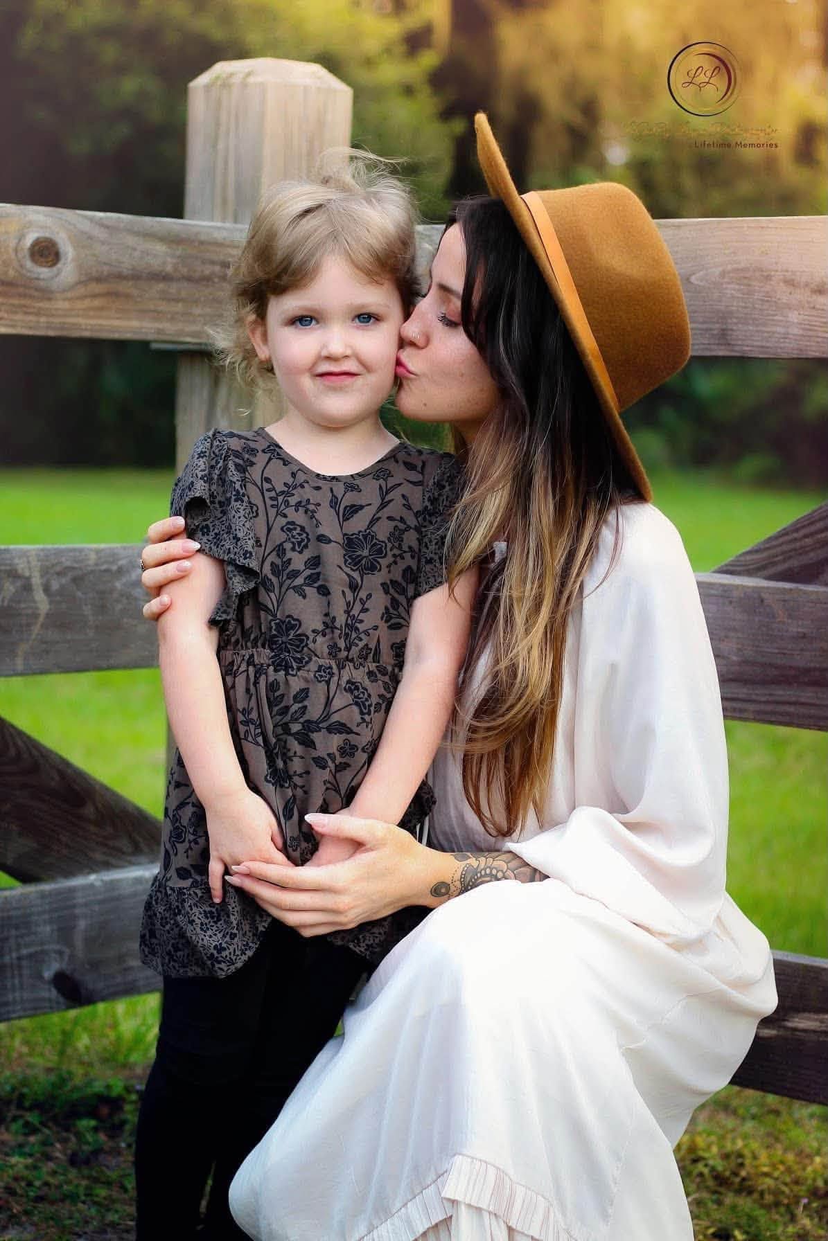 Mother kissing her young blonde daughter on the cheek near a wooden fence outdoors in Daytona Beach Florida.
