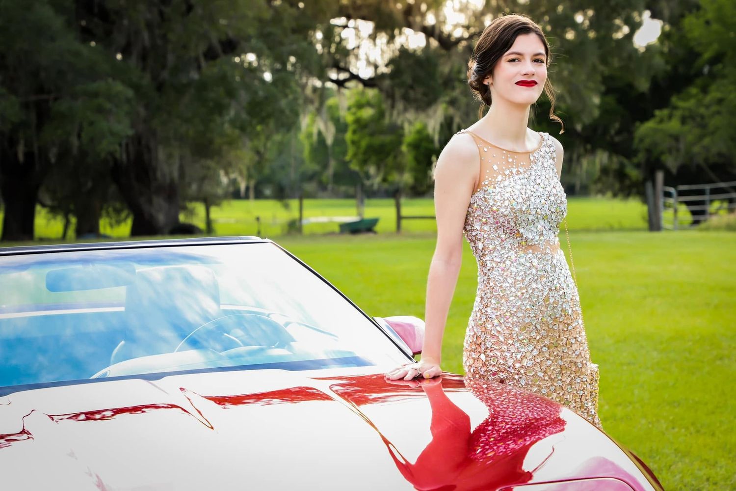 Elegant young woman in sparkling silver and red gown posing beside a classic white convertible car outdoors.