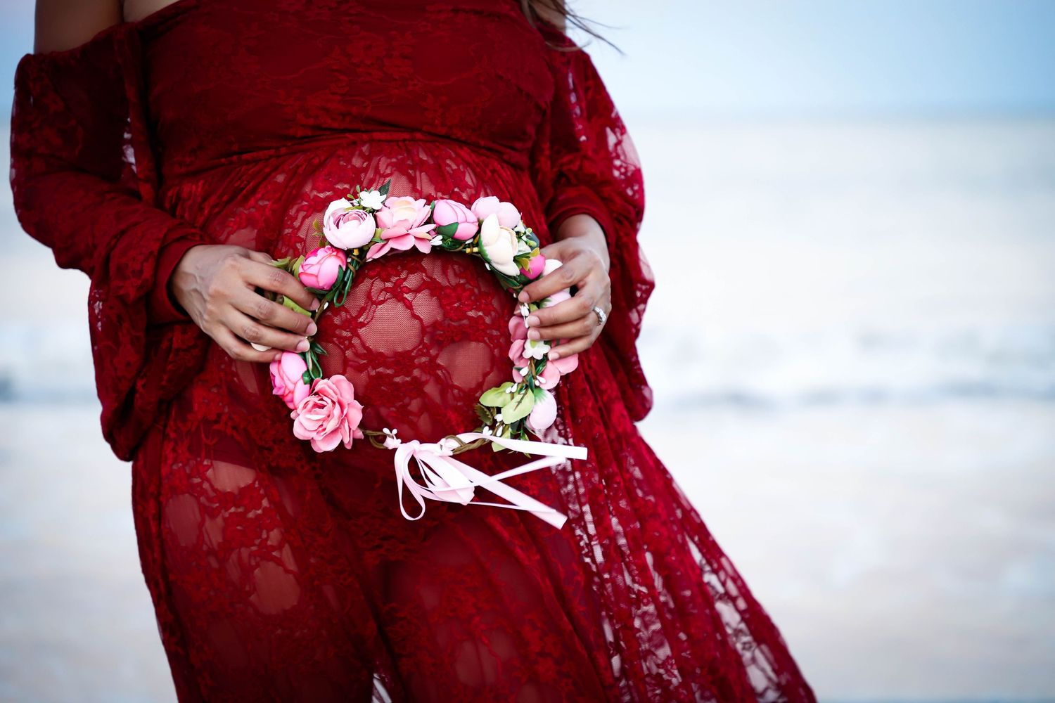 Pregnant woman in red lace dress holding floral crown over baby bump on beach.