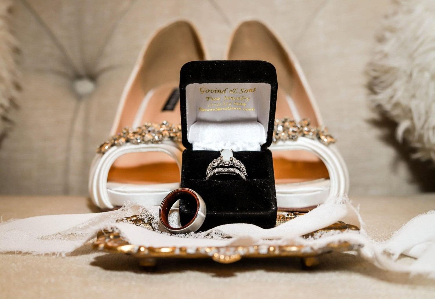 Diamond engagement ring and wedding bands displayed in black velvet box with bridal heels backdrop.