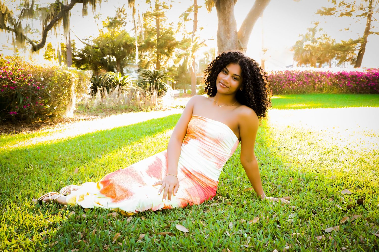 Young woman in strapless peach gradient gown sitting on green grass under sunlit trees.
