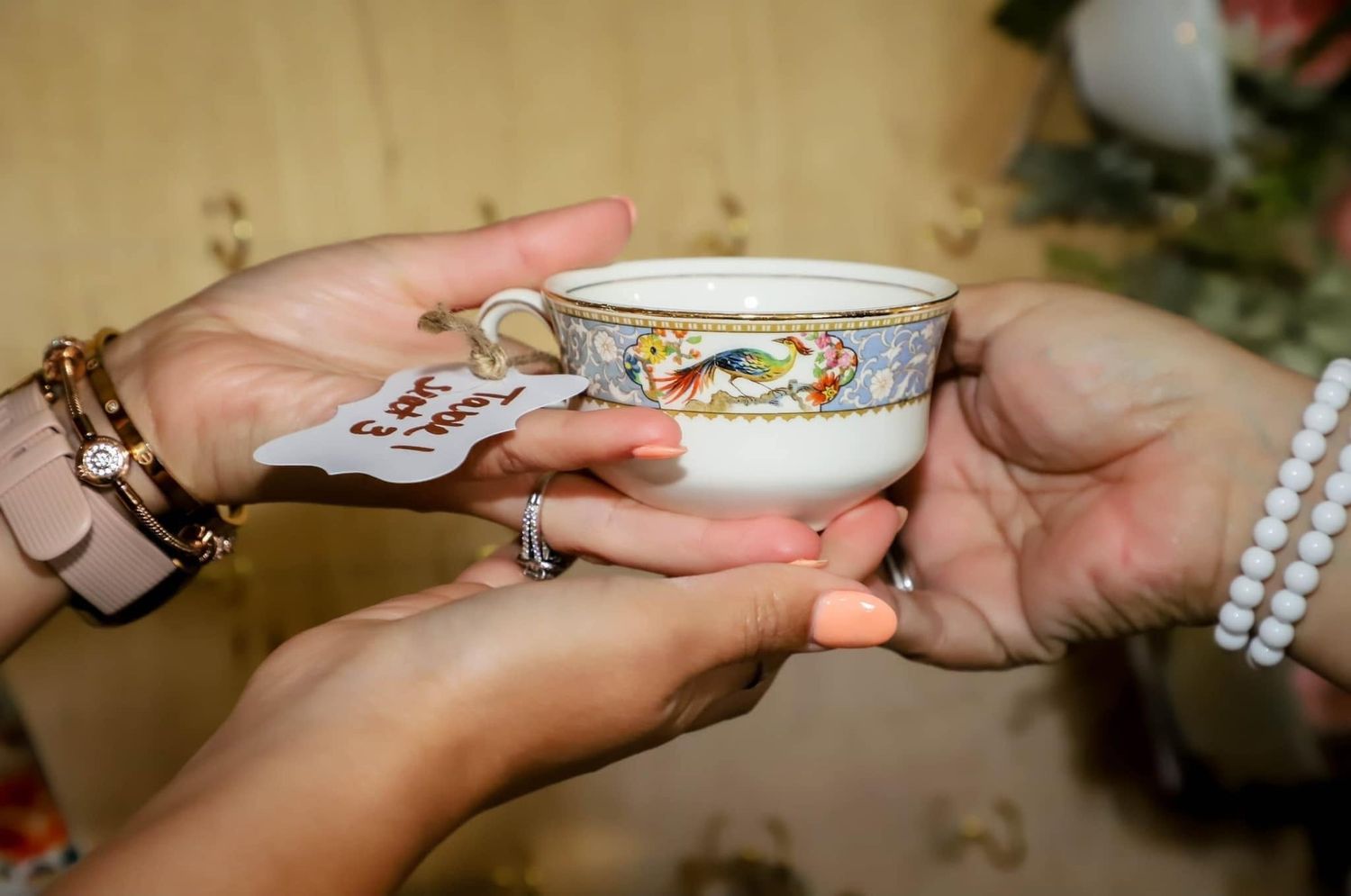Two hands gently hold an ornate floral-patterned teacup with a small tag, adorned with bracelets and rings.
