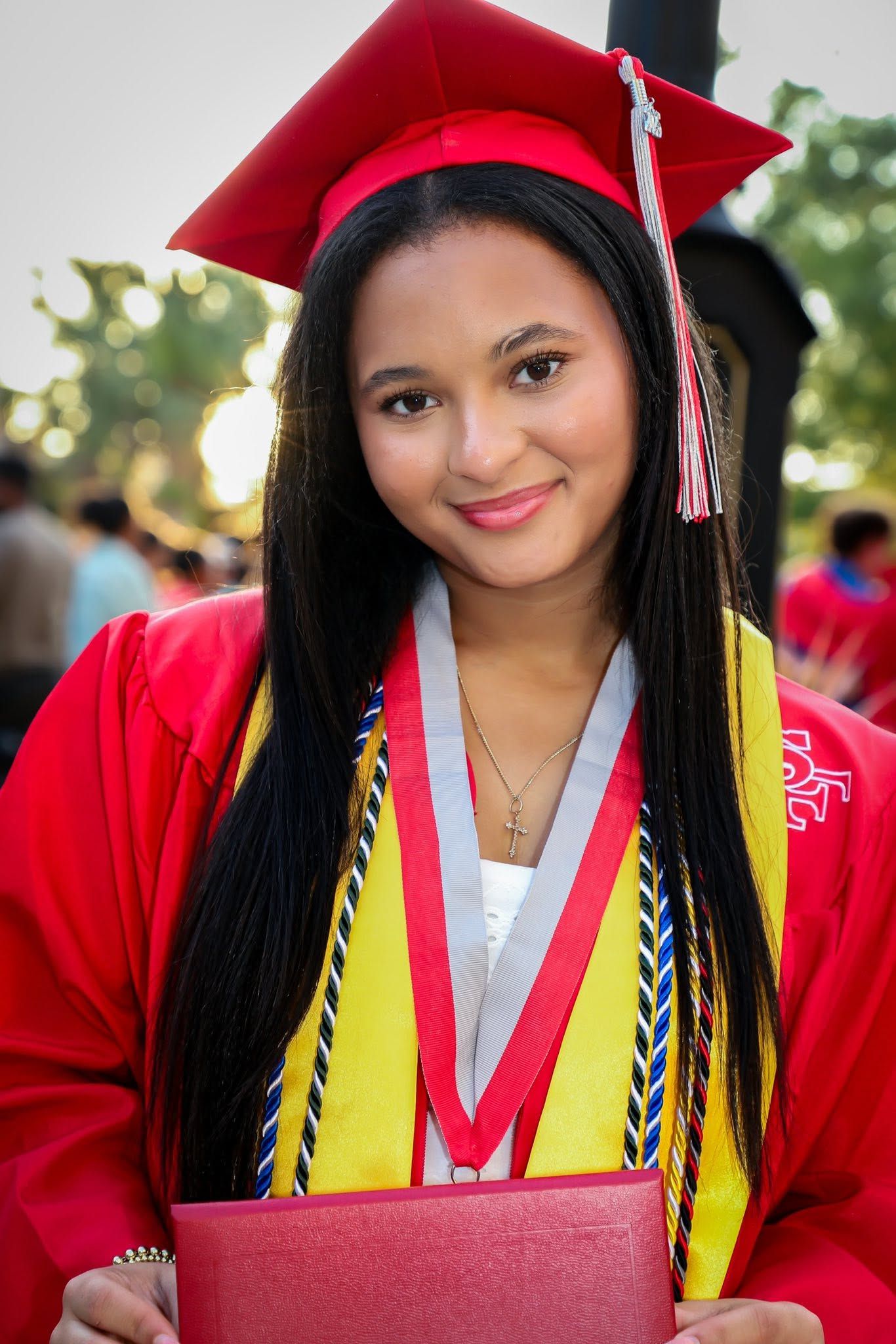 Smiling graduate in red cap and gown holding diploma at graduation ceremony.