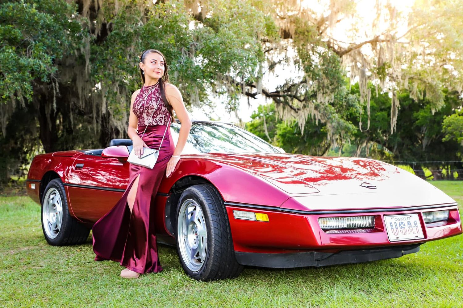 Young woman in elegant red gown posing beside classic red Corvette convertible under Spanish moss trees.