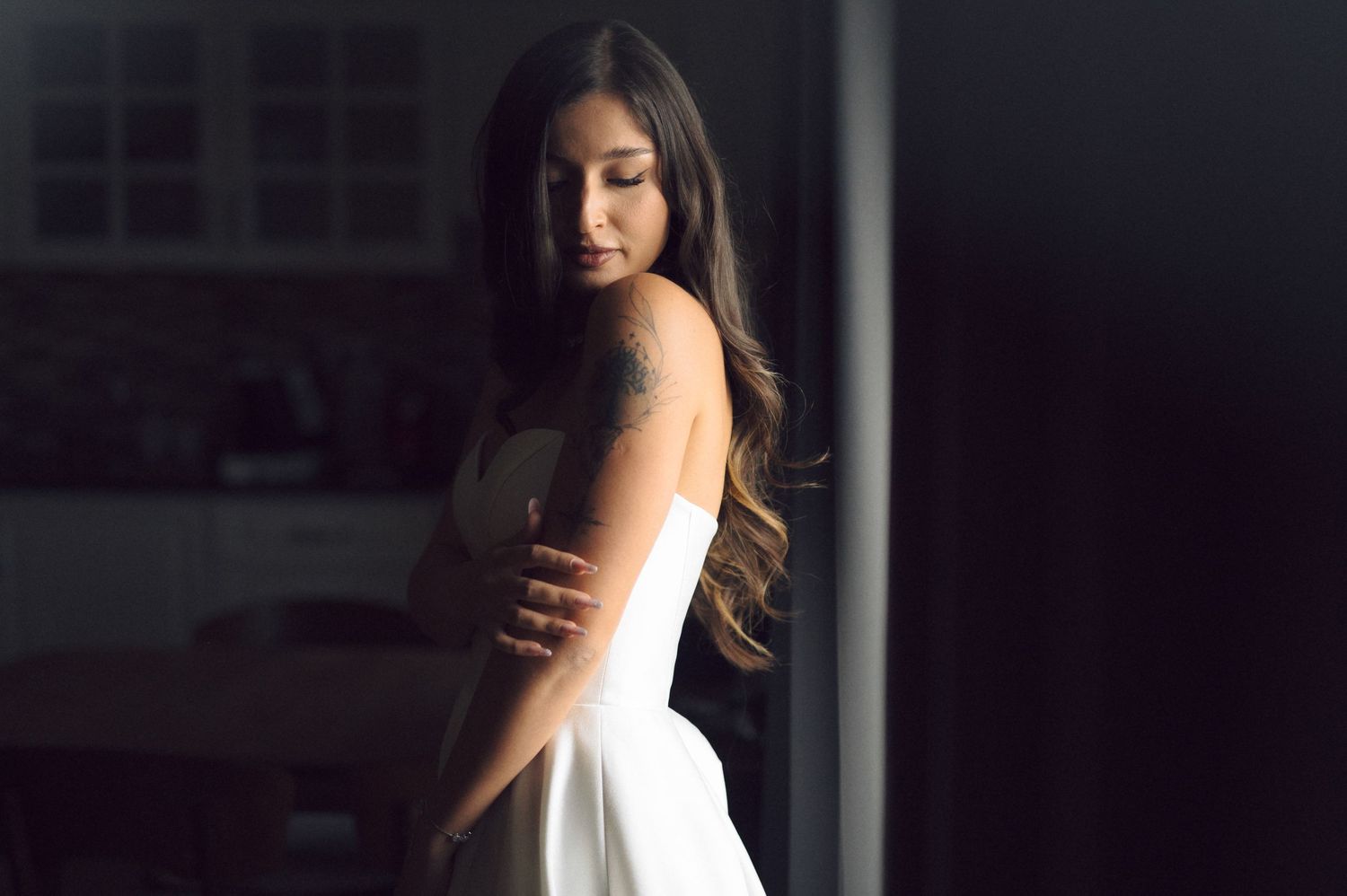 Young woman in white dress with tattoo stands in dramatic lighting, looking down thoughtfully in a modern interior.