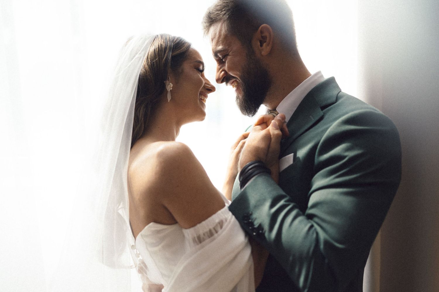 A smiling bride and groom share a romantic moment in wedding attire, with soft natural light in the background.