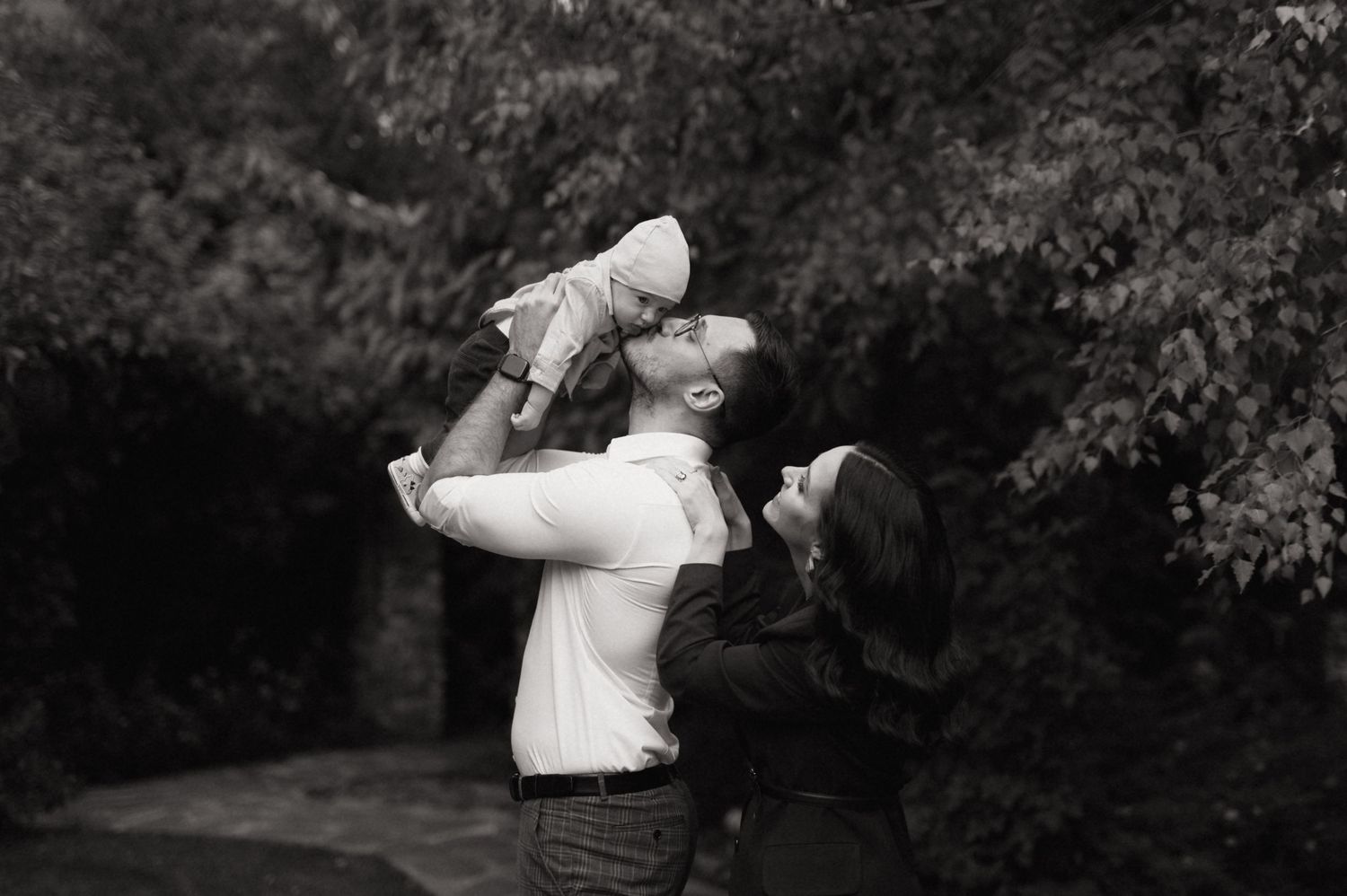 Black and white photo of parents lifting their baby up outdoors with lush green trees in the background.