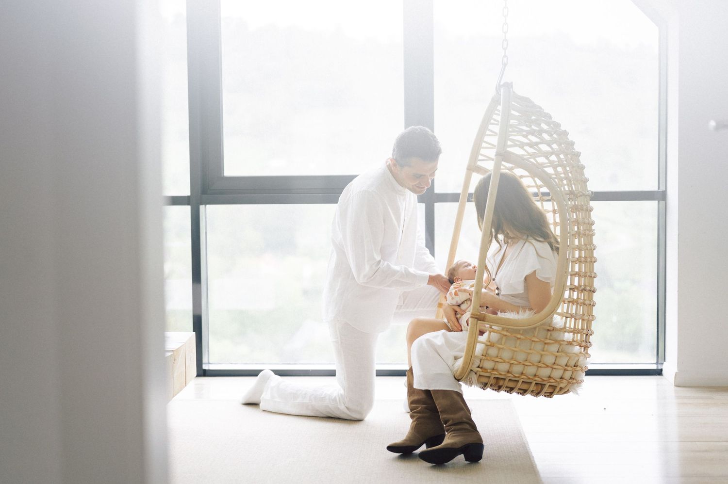 A couple in white outfits shares a tender moment near a hanging rattan chair by large windows in a bright room.