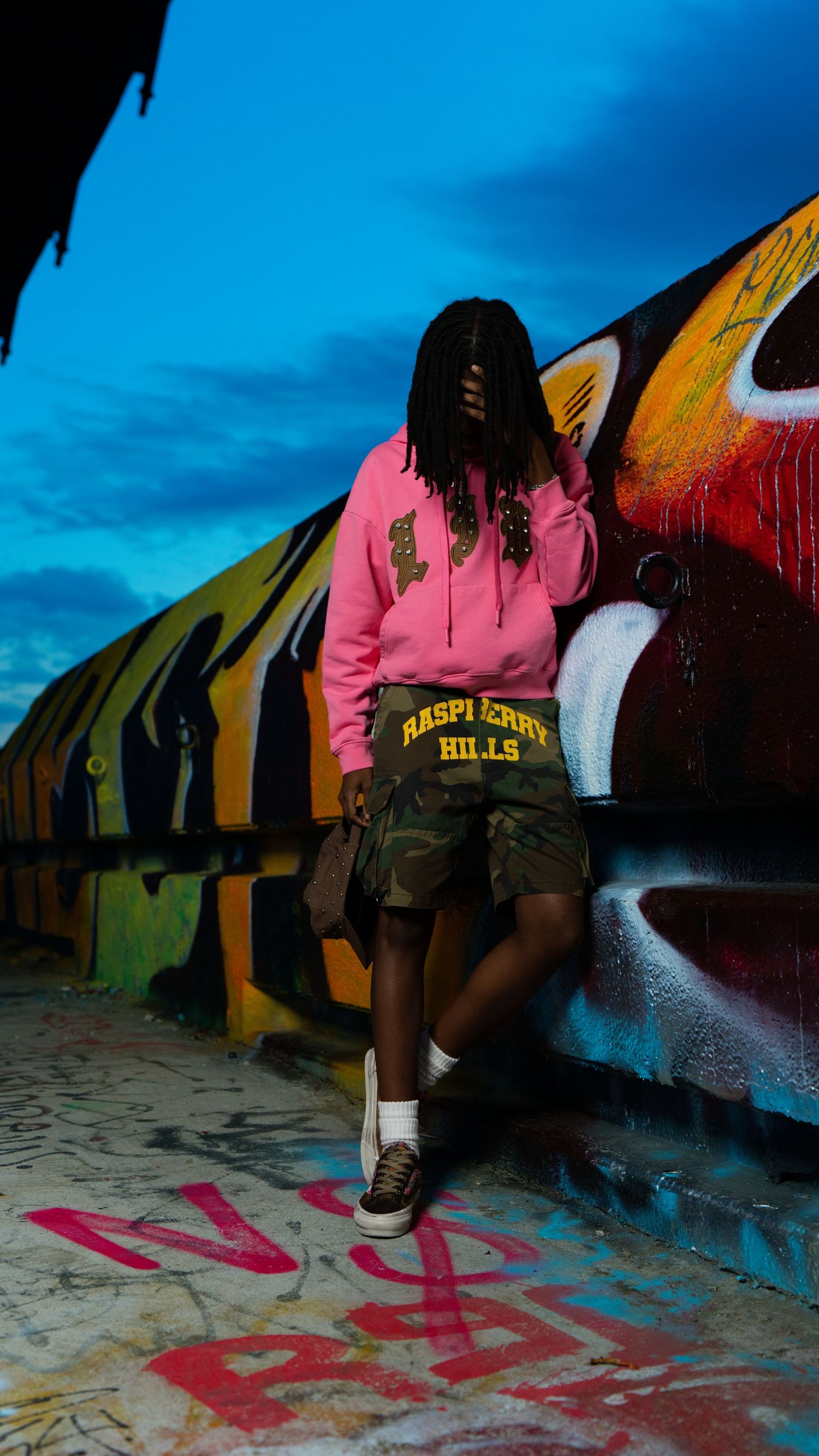 Person in pink Raspberry Hills hoodie and camo shorts poses against colorful graffiti wall at dusk.