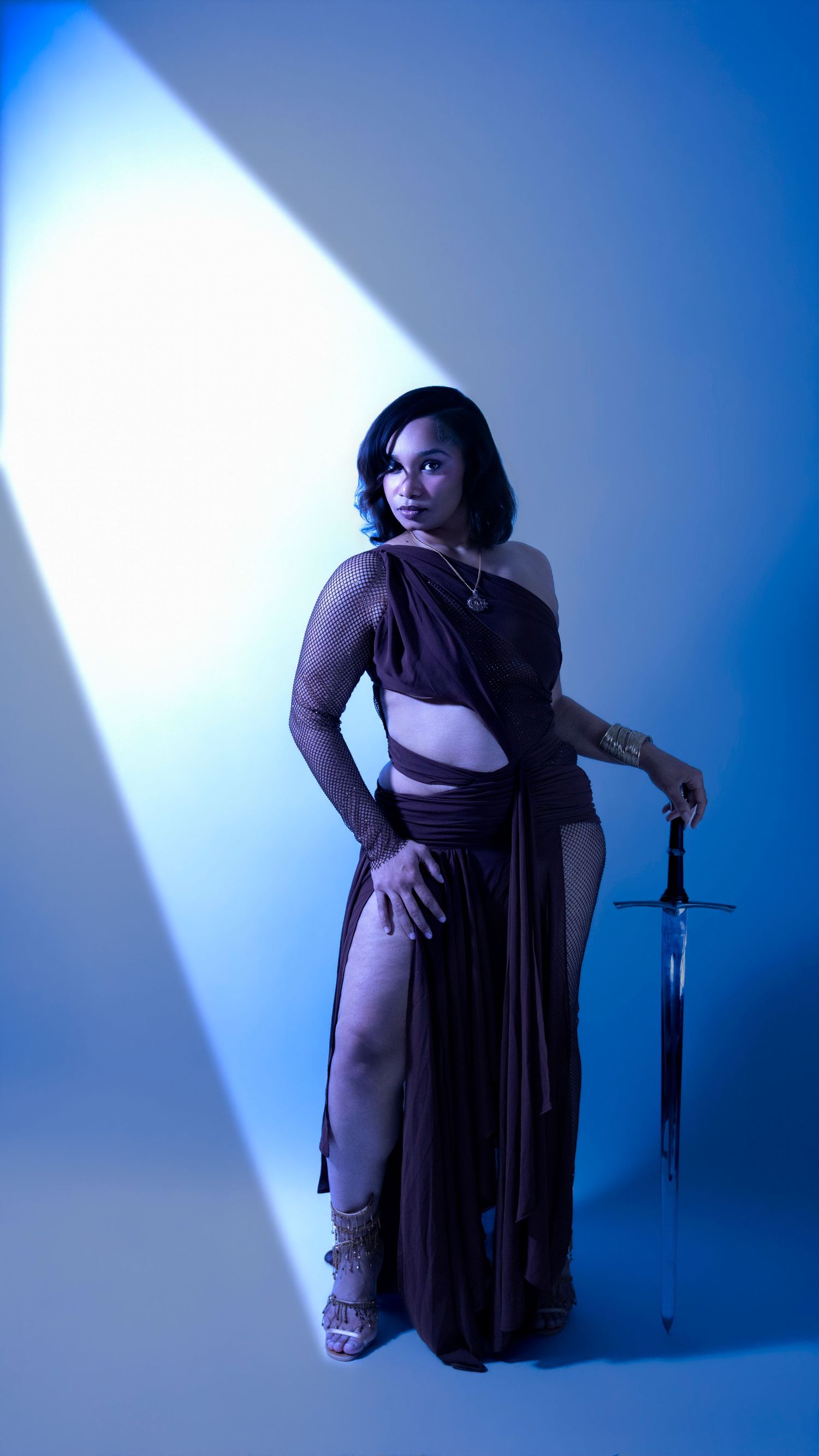 Woman in brown draped cutout gown posing with sword under dramatic blue studio lighting.