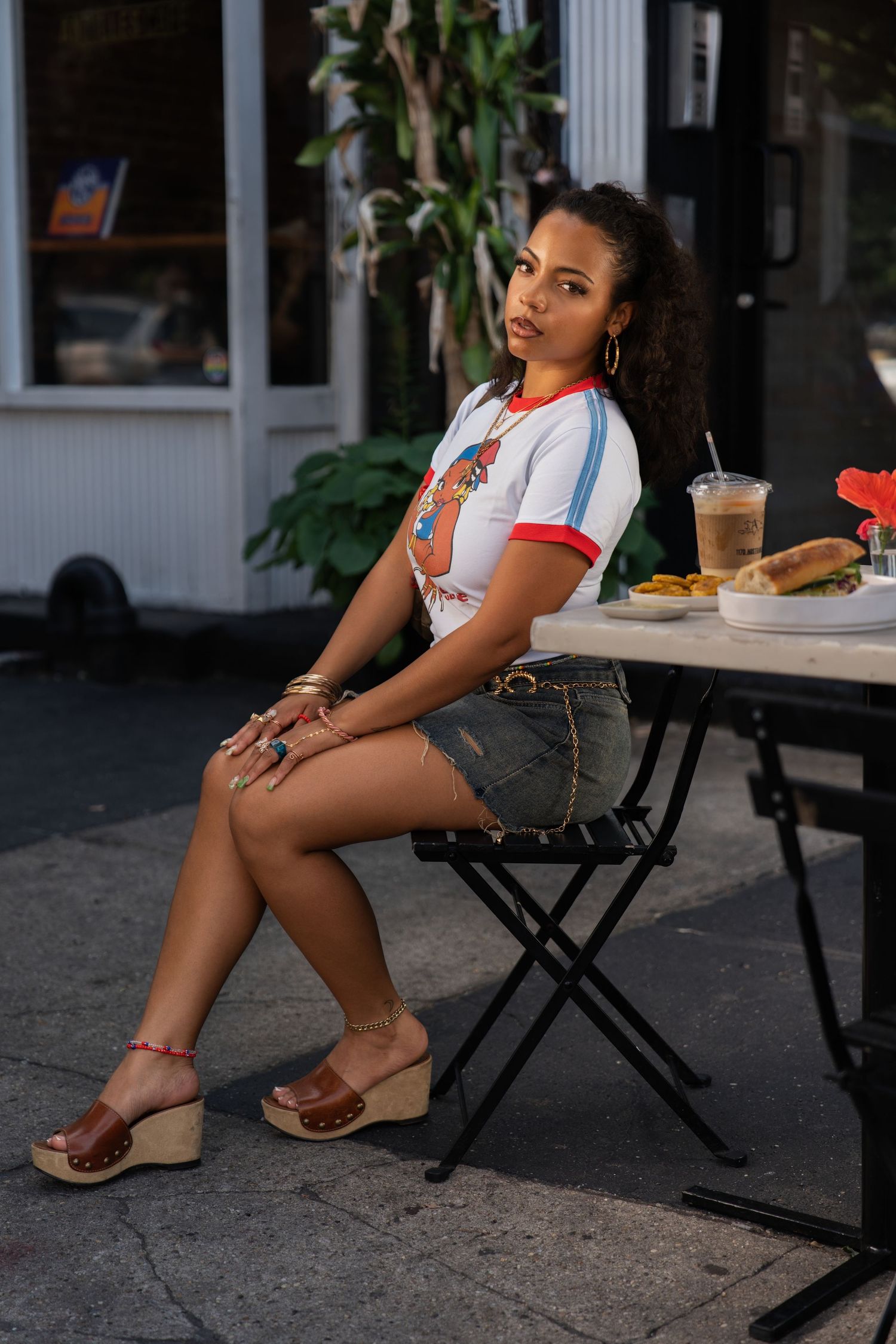 Young woman in graphic tee and denim shorts sits at outdoor cafe table with food and drinks.