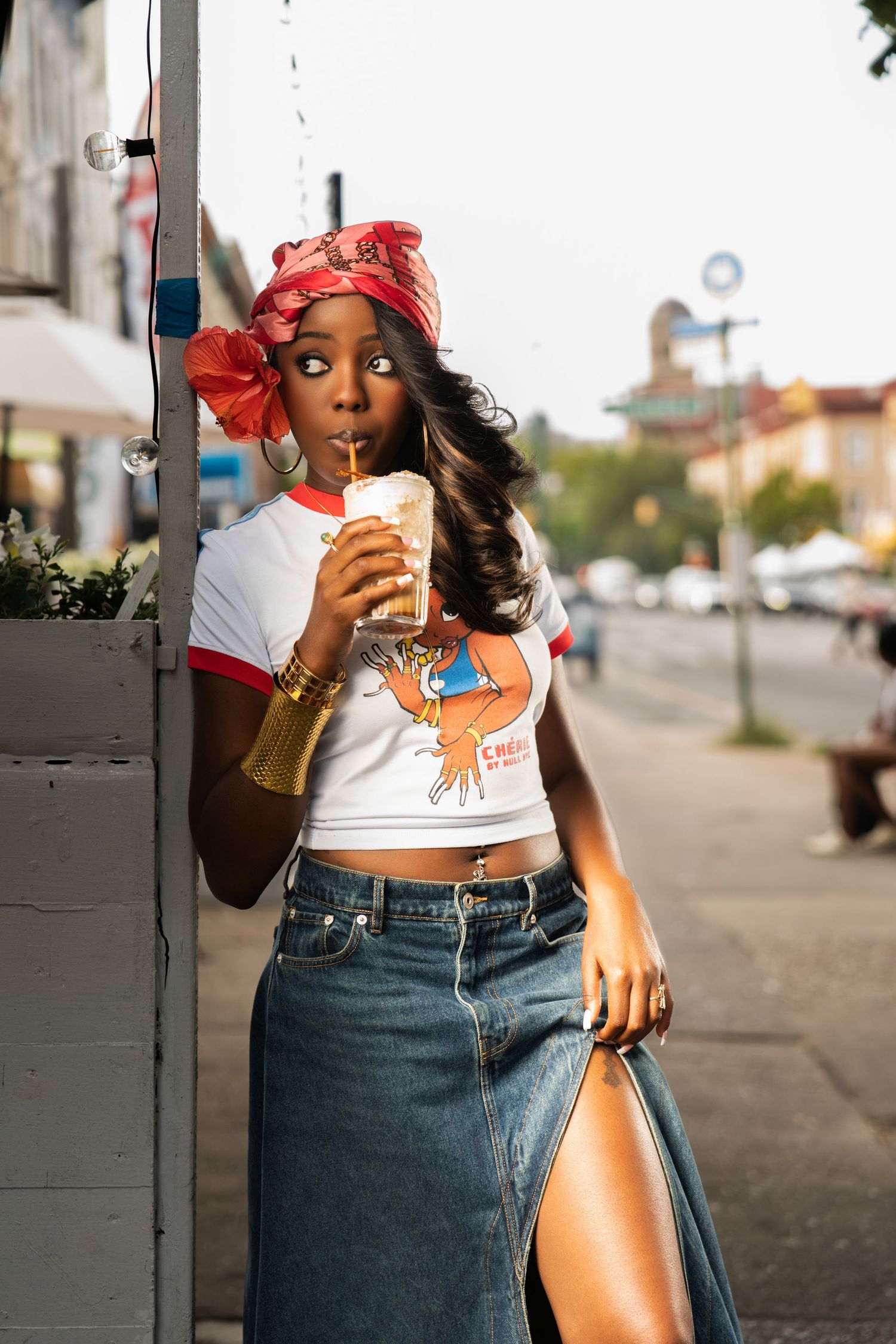 Young woman in red headscarf sips iced drink while leaning against a post on a sunny urban street.