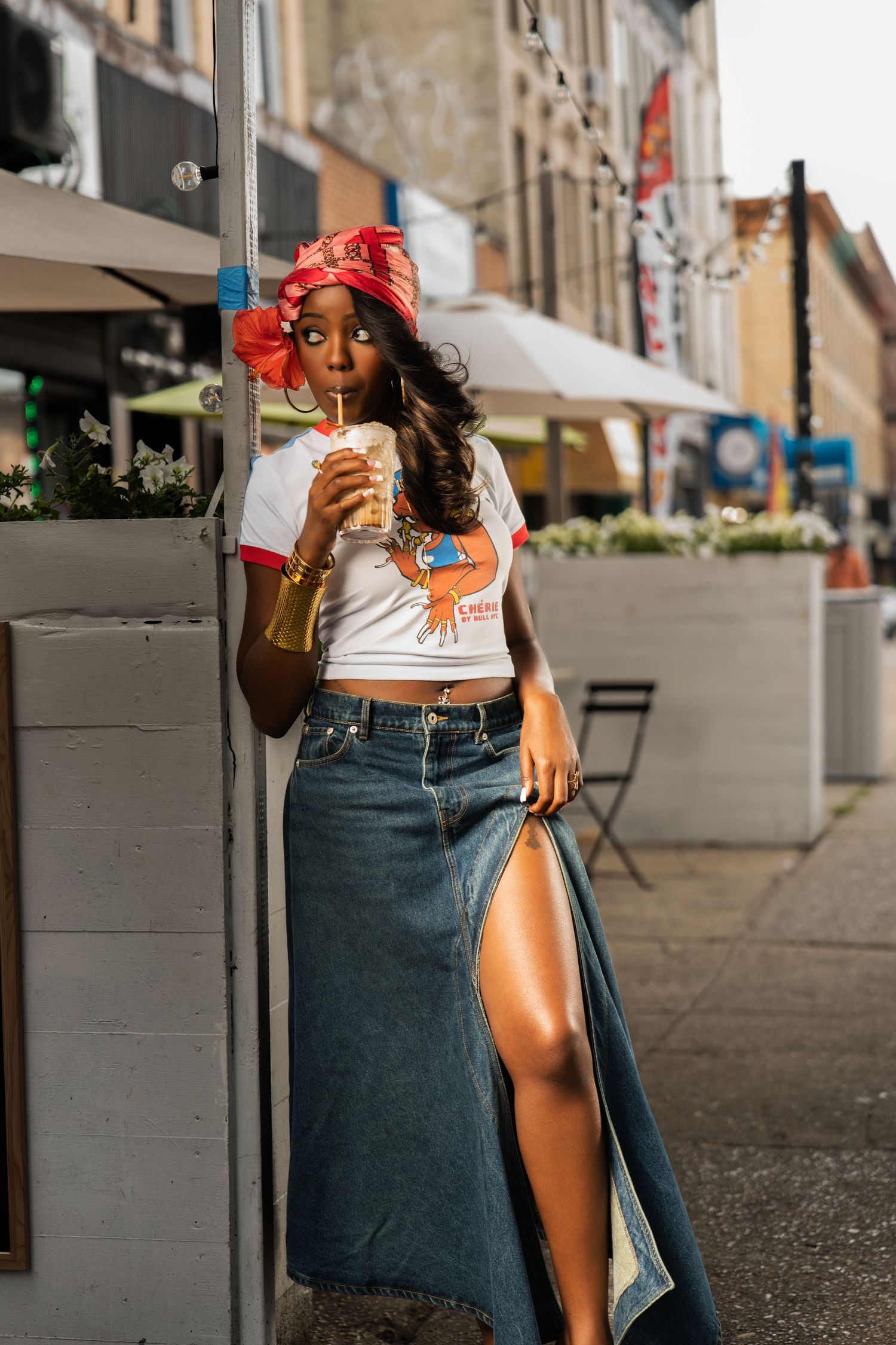 Fashionable woman in red turban and denim maxi skirt drinks iced beverage outside urban restaurant.
