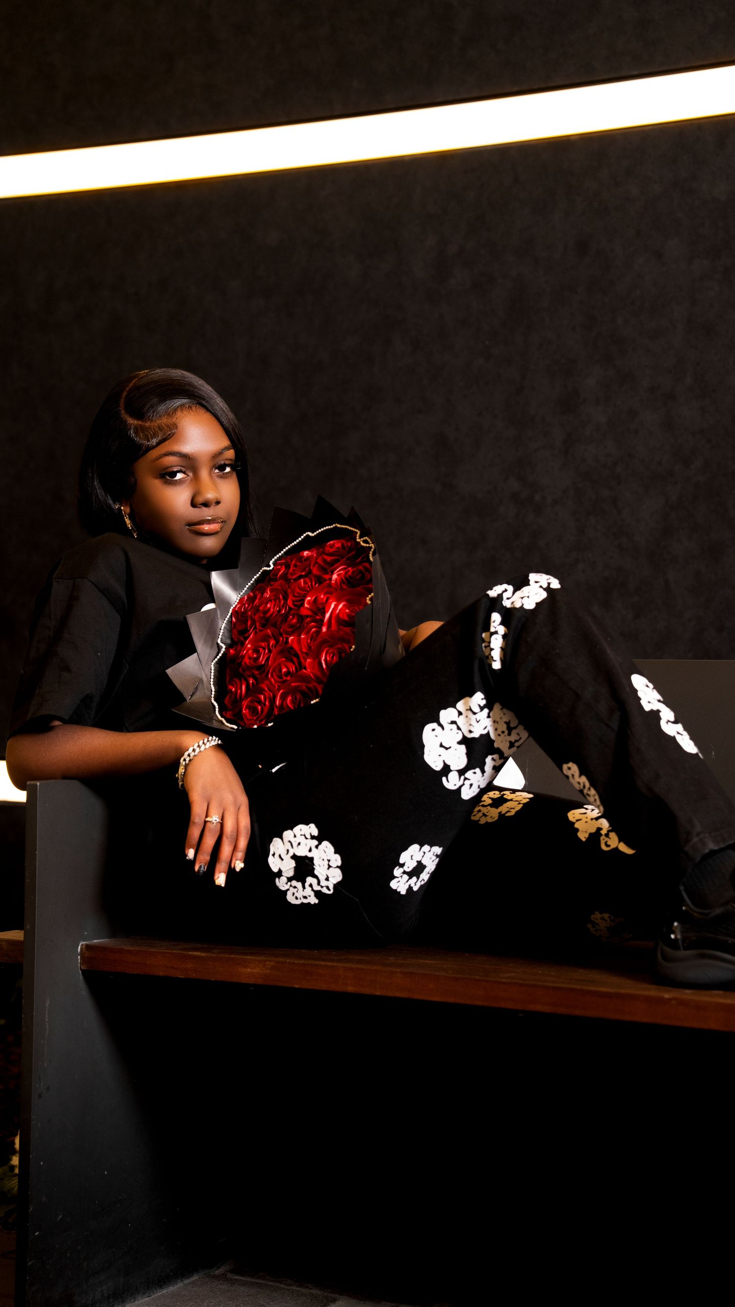 Young woman in black floral pants holds red rose bouquet while posing on bench in dark studio.