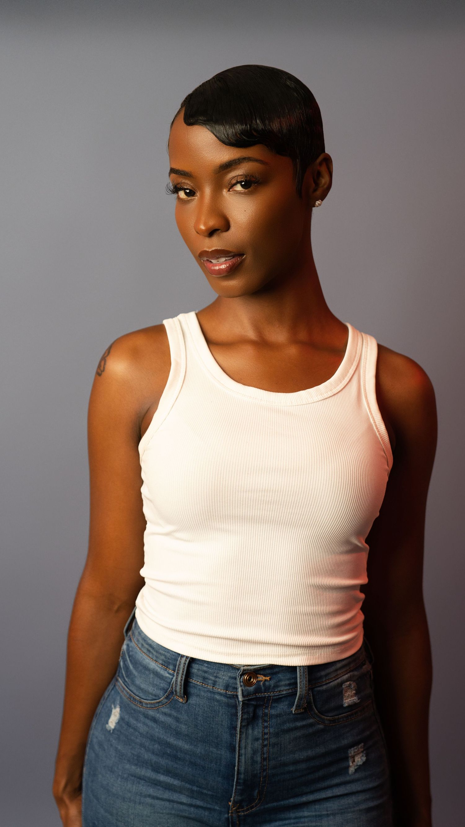 Athletic woman with short black hair wearing white tank top and distressed jeans in studio.