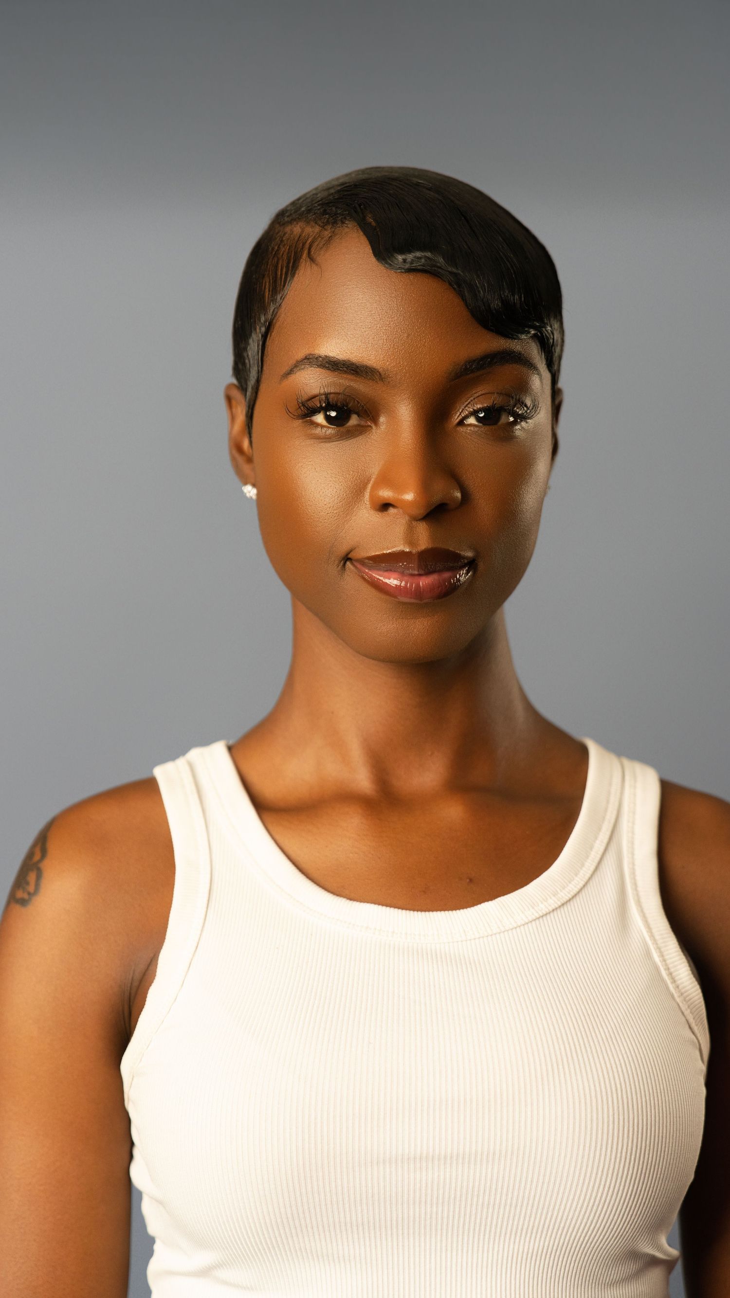 Close-up portrait of woman with sleek short hair in white tank top against gray background.