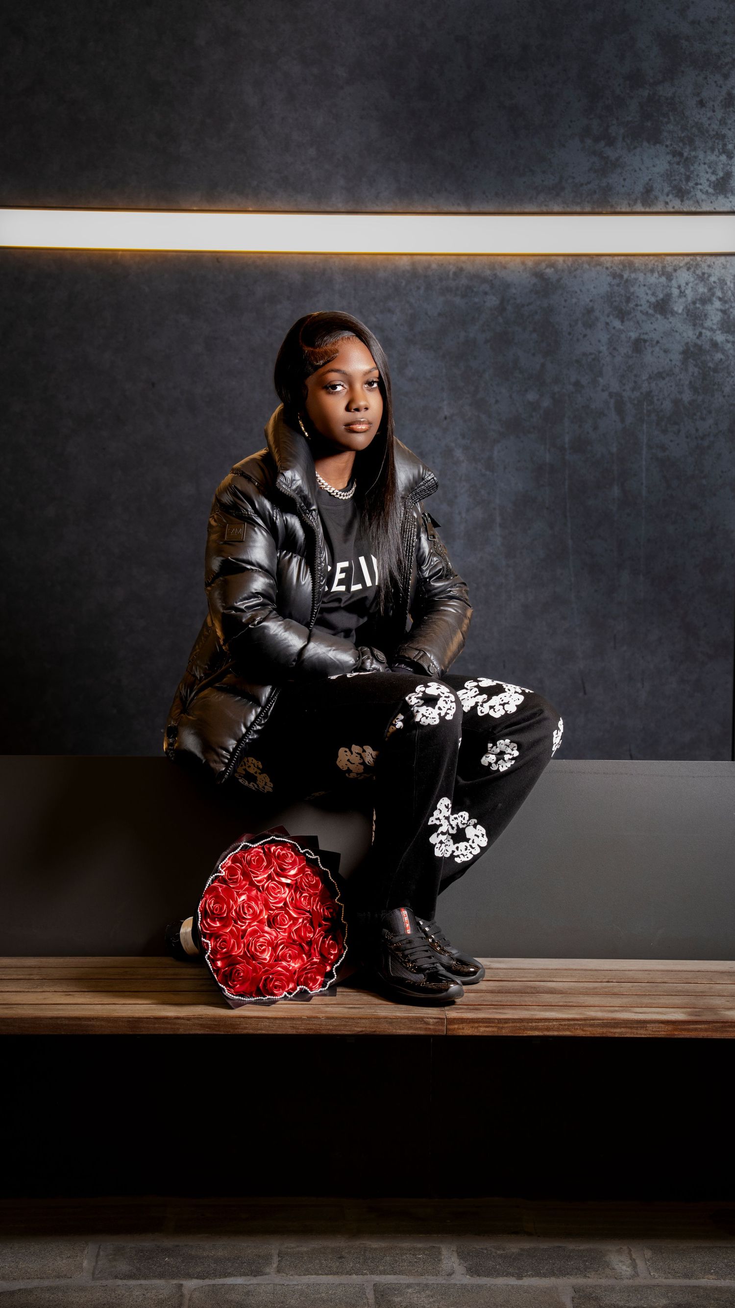 Young woman in black puffer jacket and floral pants poses with red roses on wooden bench.