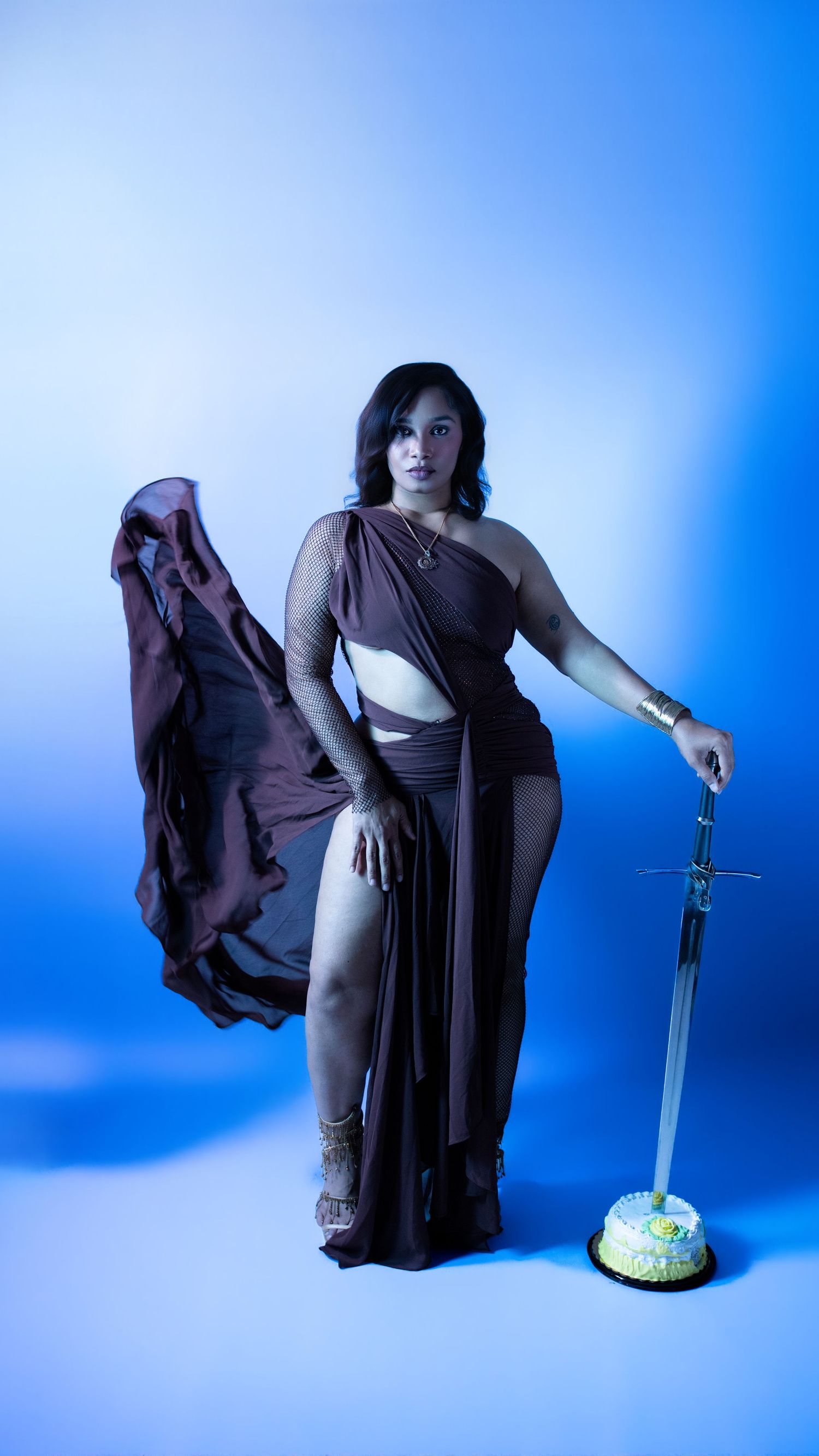 Woman in flowing brown cutout dress holding sword beside birthday cake on blue background.