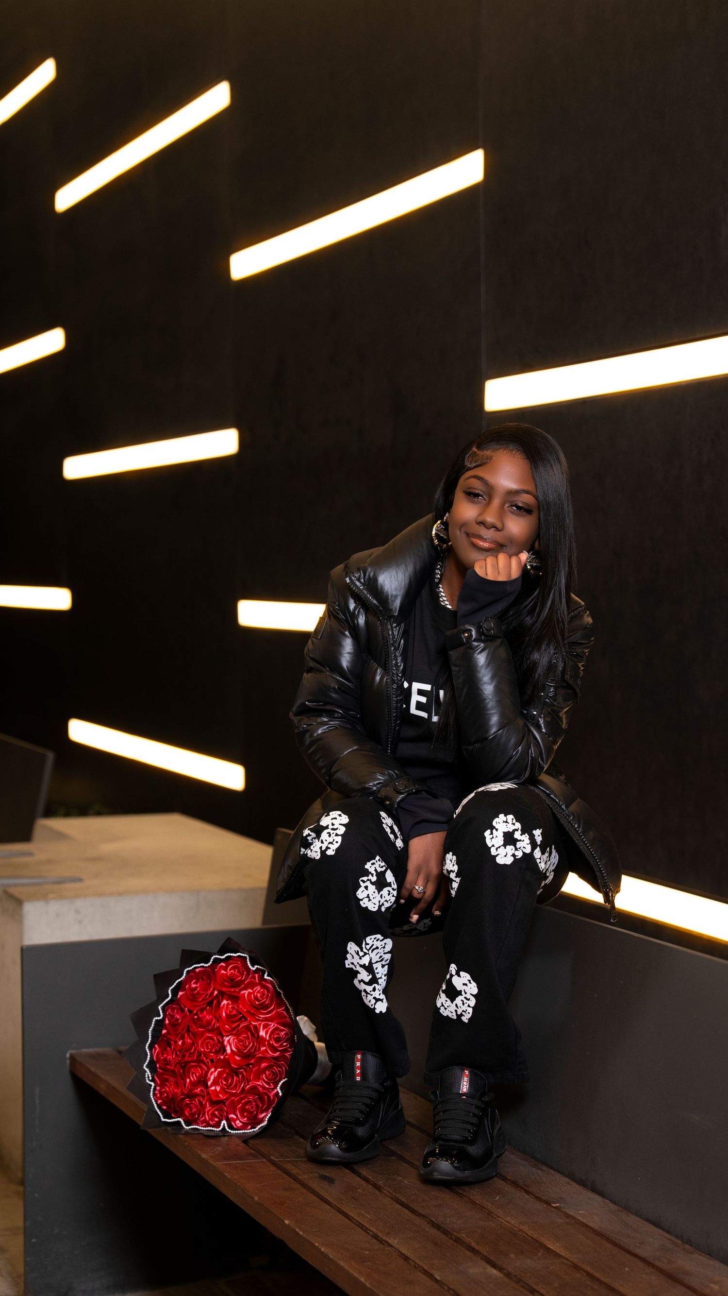 Smiling young woman in black leather jacket sits on bench with red rose bouquet against lit wall.