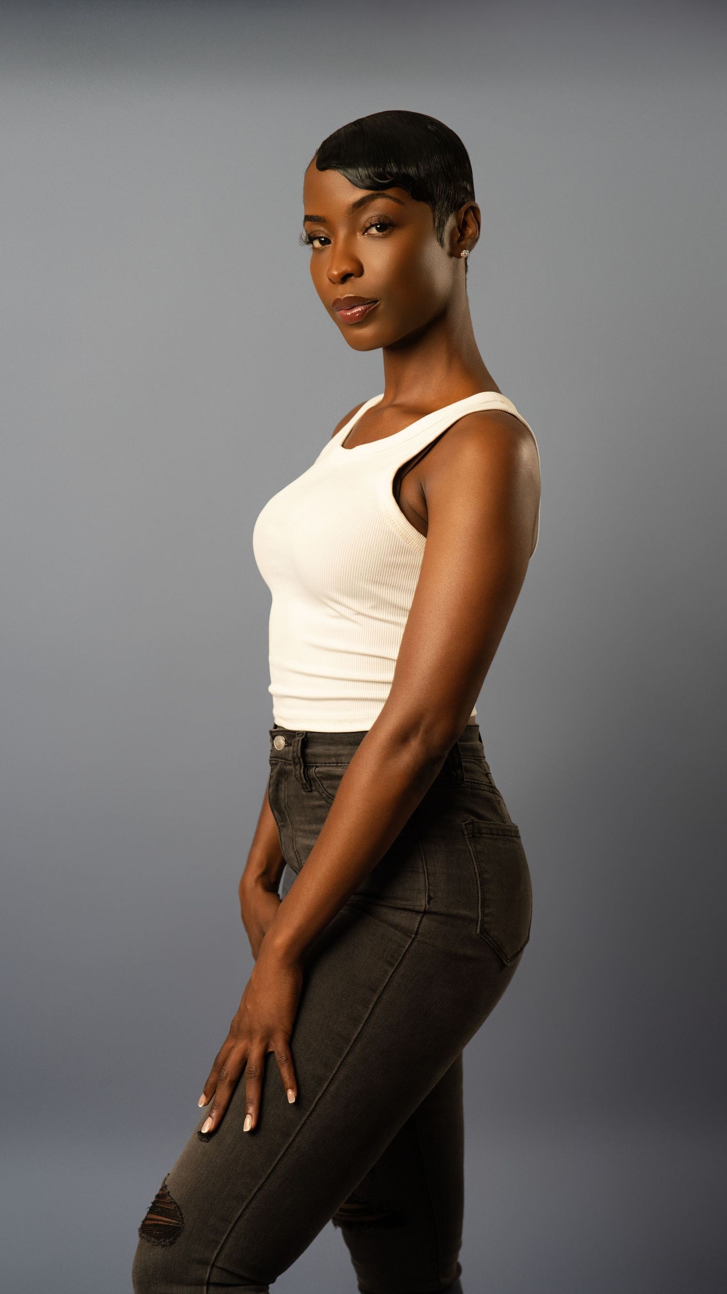 Woman in white tank top and dark jeans poses in side profile against neutral gray backdrop.